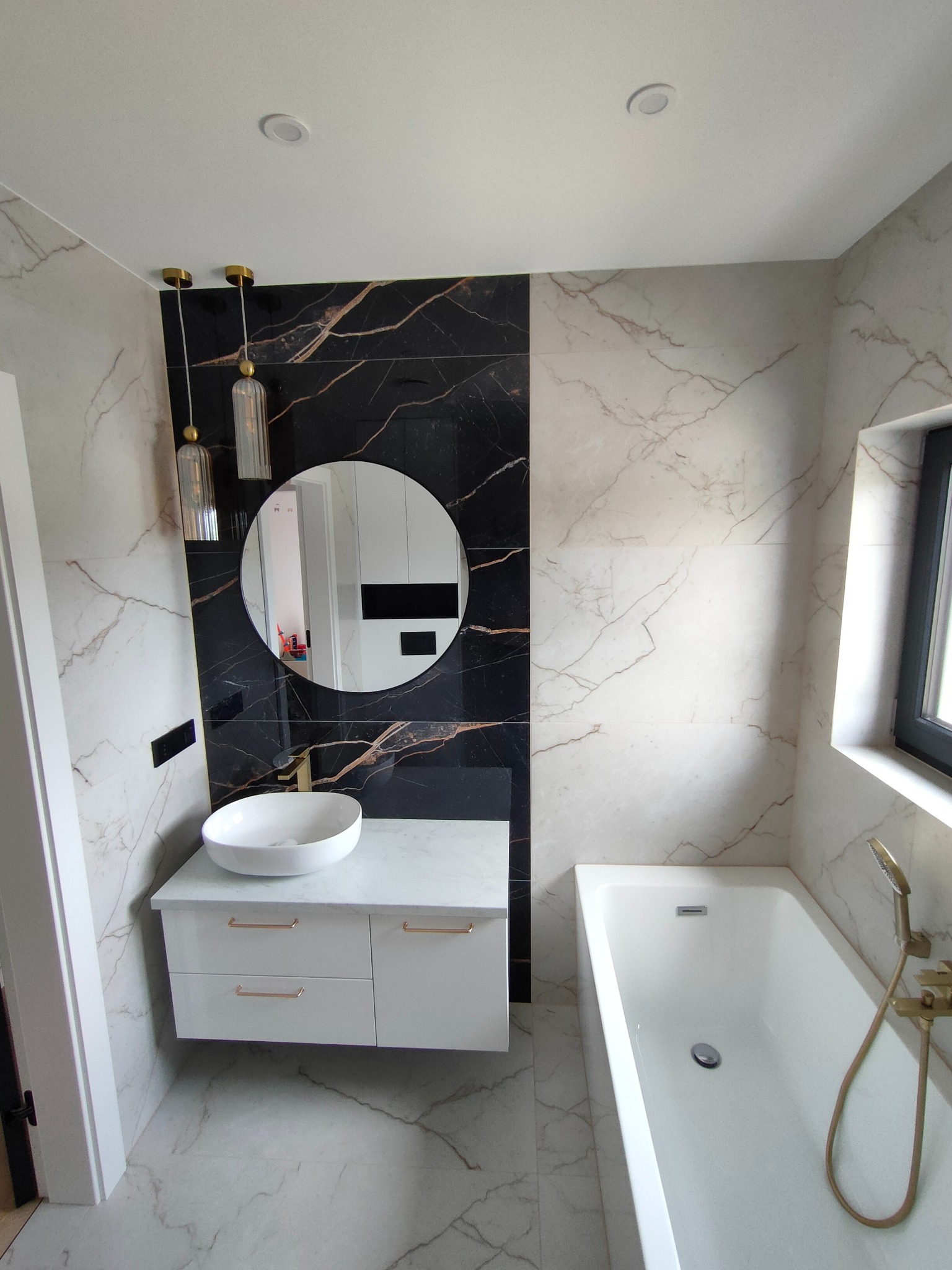 Elegancka łazienka z marmurowymi płytkami, okrągłym lustrem, wiszącą szafką z umywalką i złotą armaturą. Minimalistyczny design z akcentami czerni i złota.