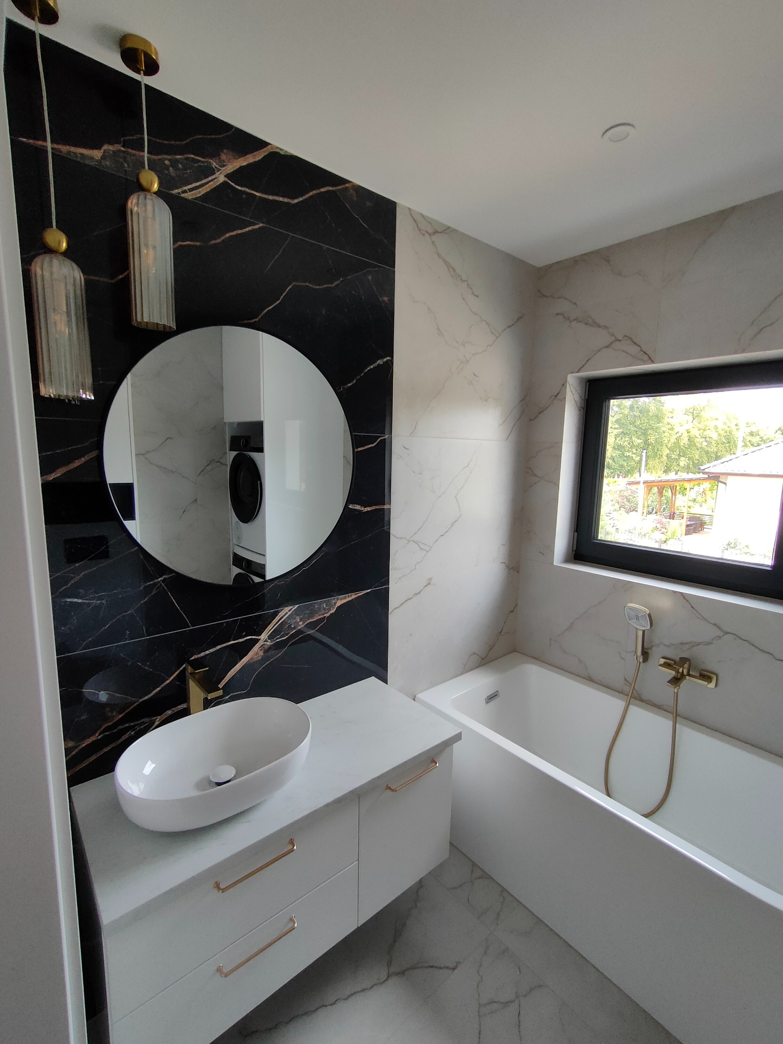 Elegancka łazienka: czarne i białe marmurowe płytki, owalna umywalka, złota armatura, okrągłe lustro odbijające pralkę. Nowoczesne lampy wiszące.