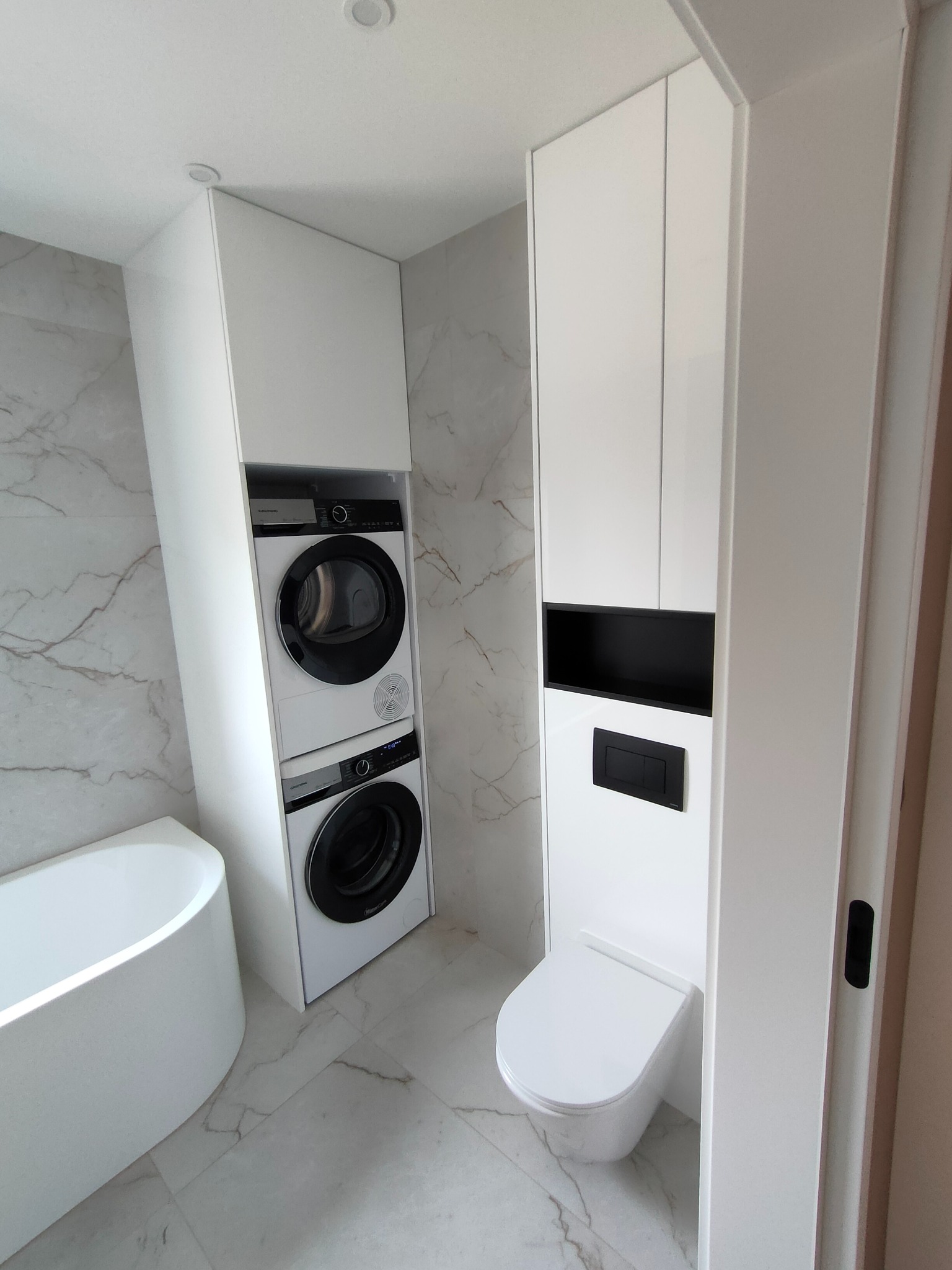 Nowoczesna łazienka z białą armaturą i szafkami w zabudowie, marmurowe płytki, pralka i suszarka w kolumnie. Minimalistyczny design.