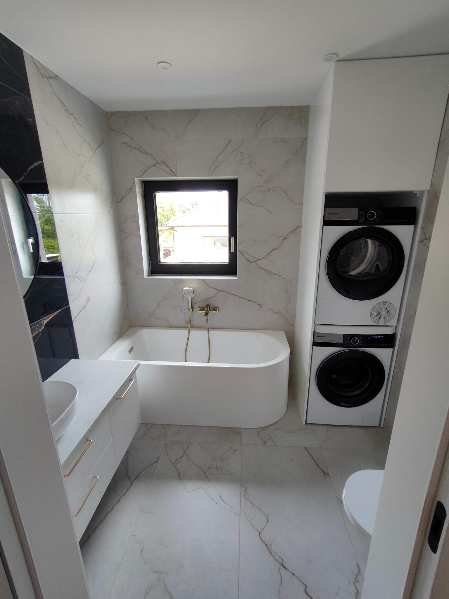 Nowoczesna łazienka z marmurowymi płytkami, białą wanną, umywalką z szafką i pralko-suszarką w zabudowie. Okno z czarną ramą. Armatura w kolorze złotym.
