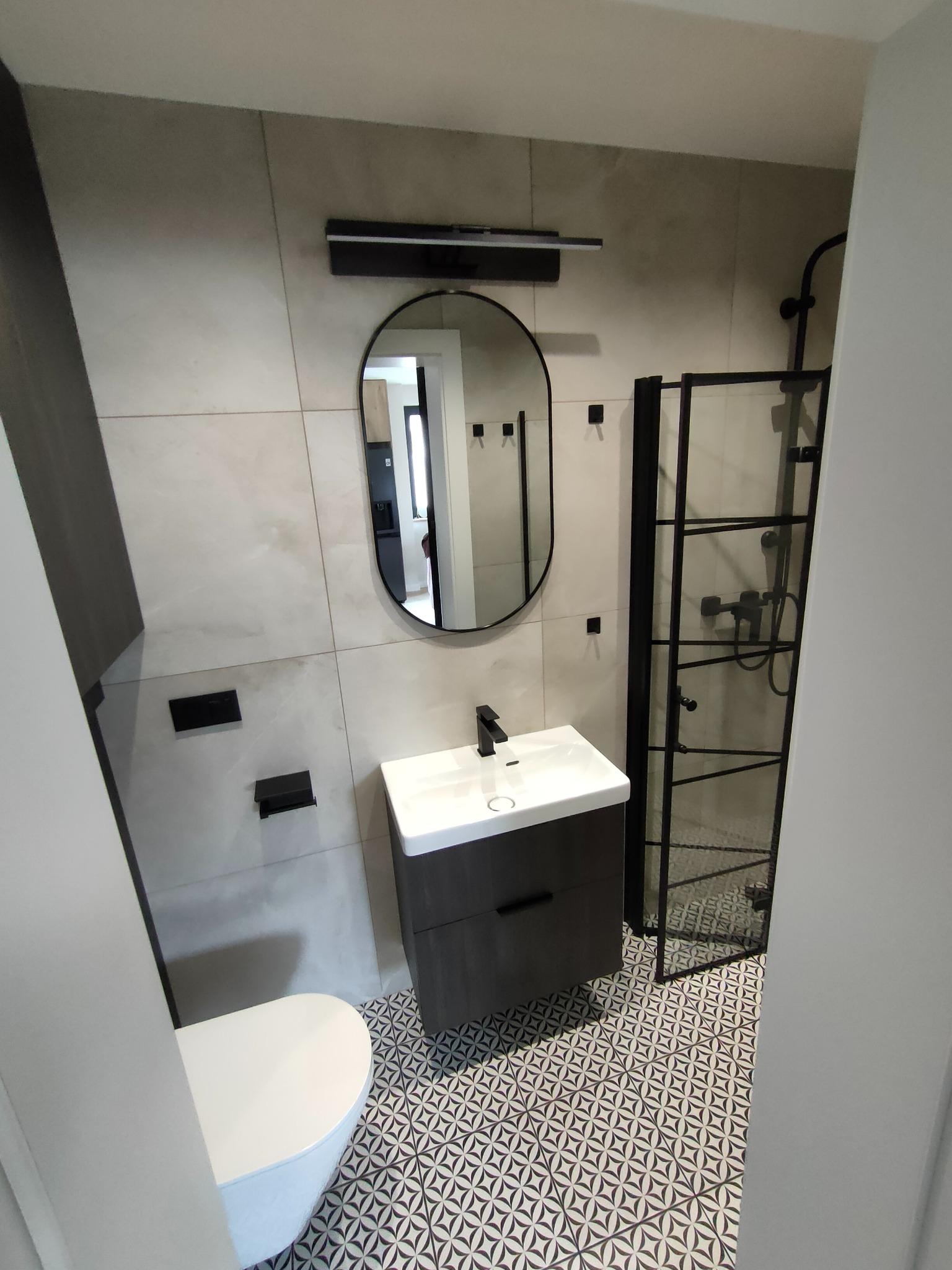 Nowoczesna łazienka po remoncie: czarne akcenty, geometryczne płytki podłogowe, owalne lustro i kabina prysznicowa typu walk-in.