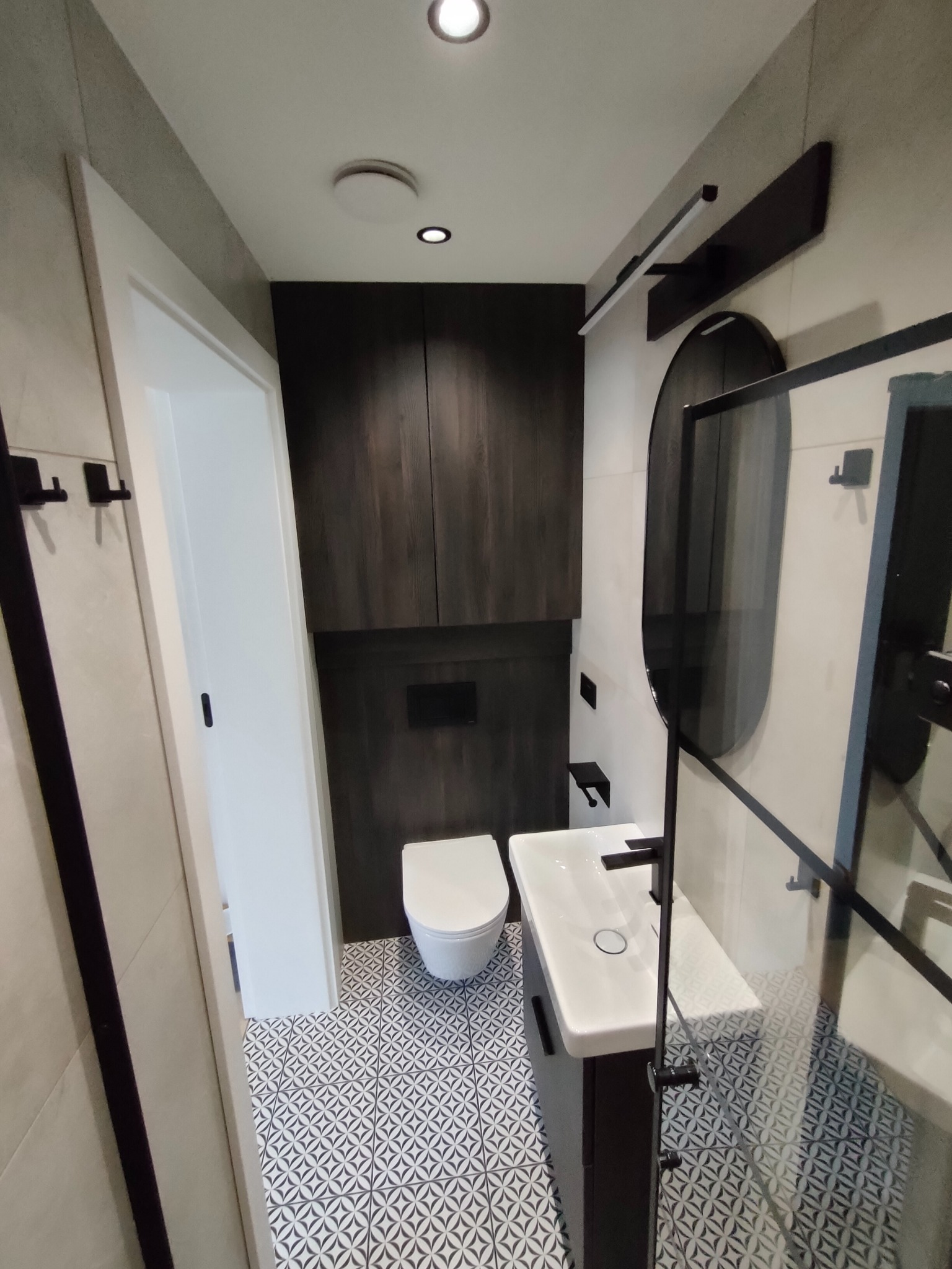 Nowoczesna łazienka z ciemnymi szafkami, owalnym lustrem i kabiną prysznicową typu walk-in. Wzór płytek podłogowych w geometryczne motywy.