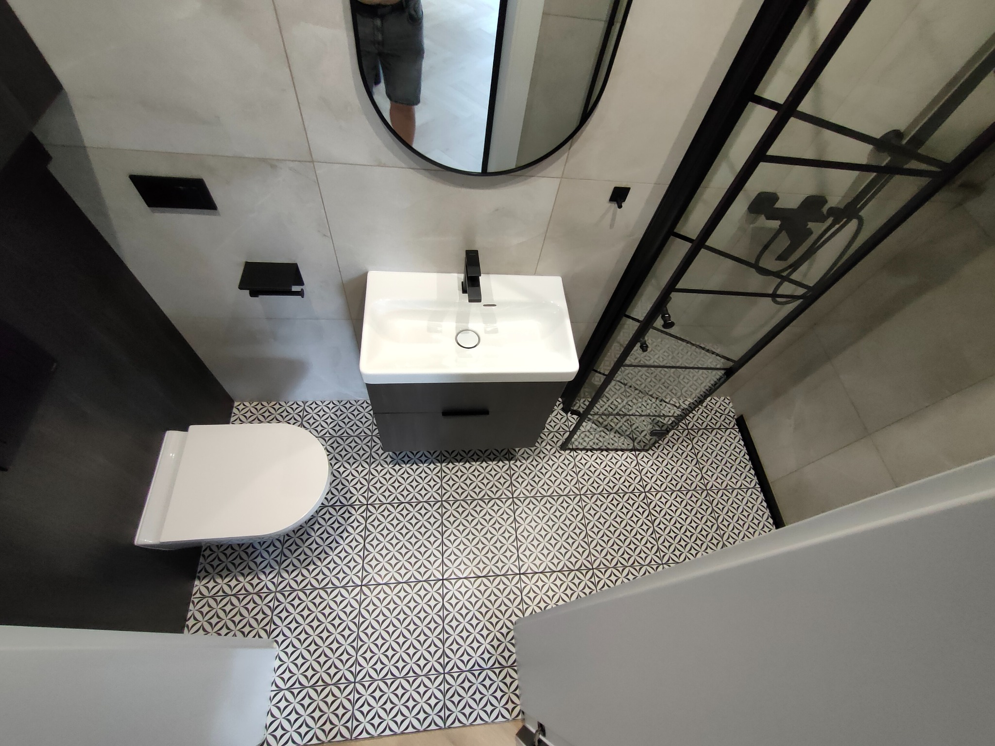 Nowoczesna łazienka po remoncie: biała ceramika, czarne akcenty, wzorzysta podłoga. Umywalka z szafką, toaleta i kabina prysznicowa z czarną armaturą.