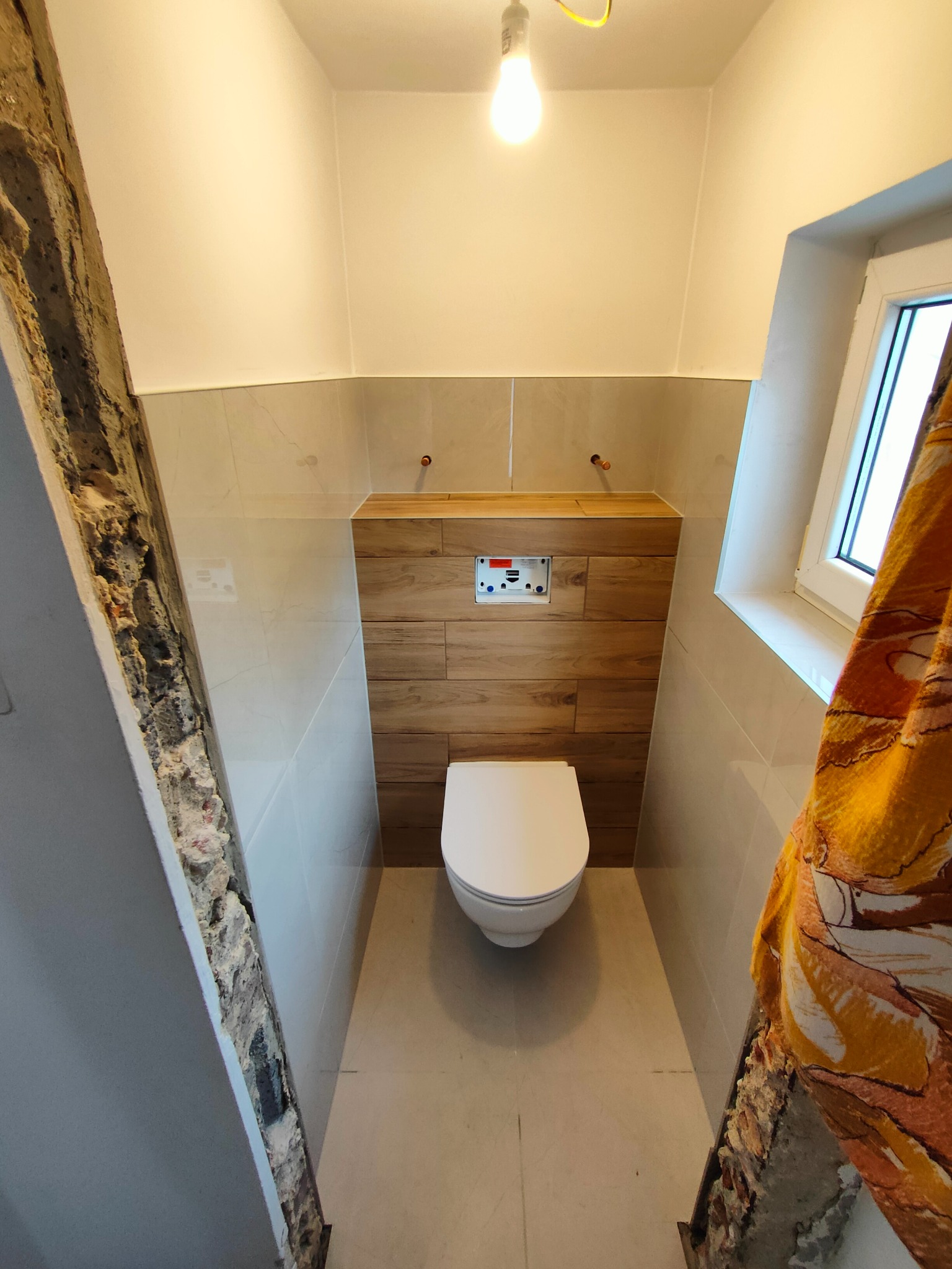 Wykończona toaleta z podwieszaną muszlą klozetową, obudowaną drewnopodobnymi płytkami, jasnymi płytkami na ścianach i widocznymi fragmentami surowej ściany.