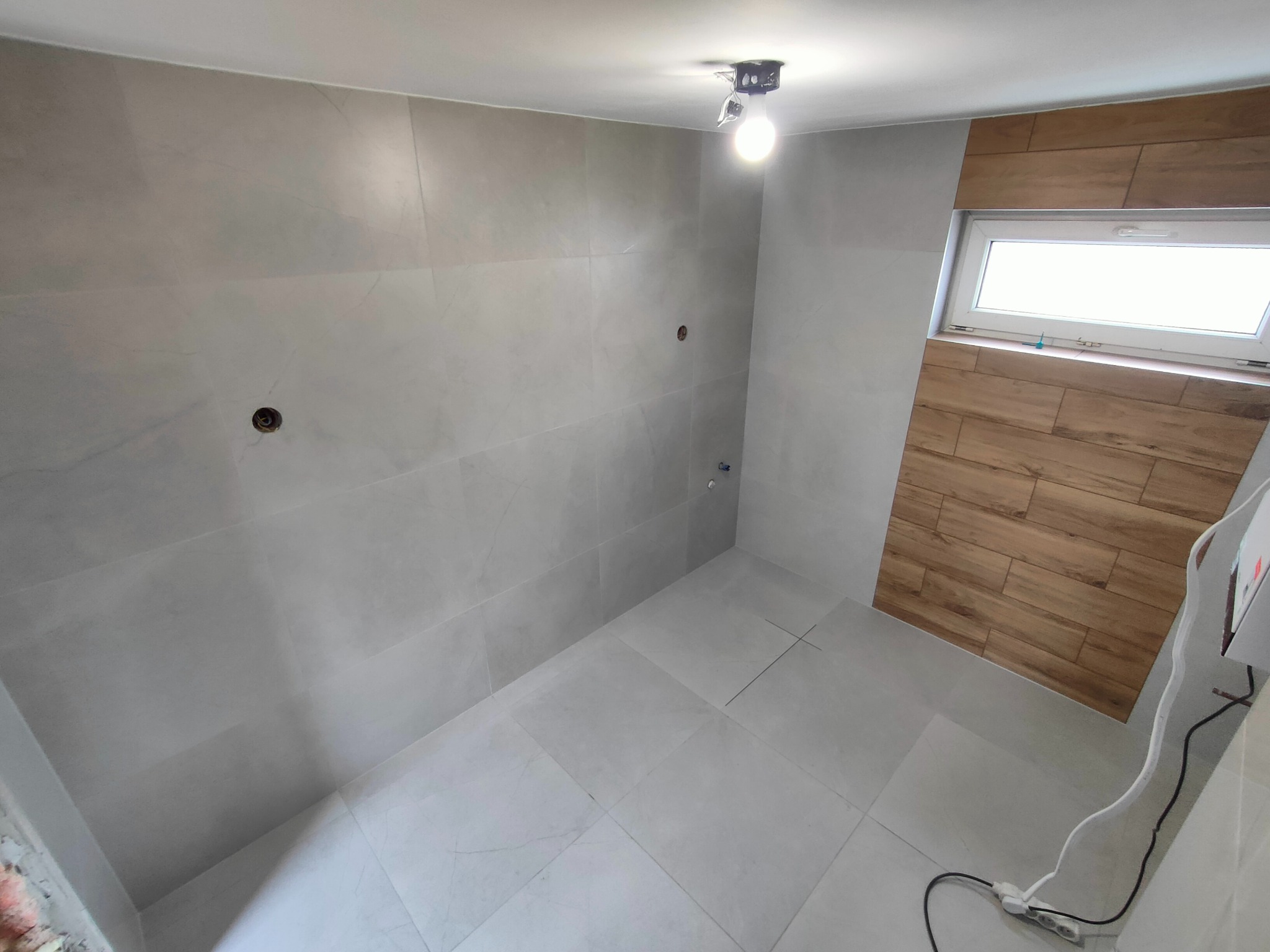 Ukończona łazienka z szarymi płytkami wielkoformatowymi na ścianach i podłodze, drewnopodobne płytki wokół okna, surowa żarówka. Widok na narożnik.