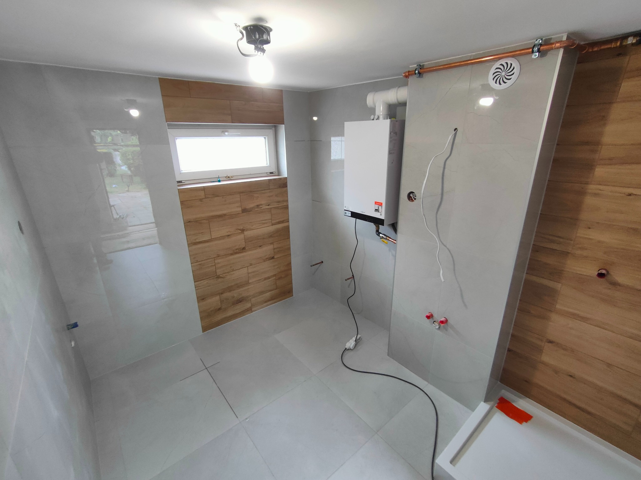 Wykończona łazienka z szarymi i drewnopodobnymi płytkami. Widoczny piec gazowy, wentylacja i instalacja. Ujęcie z bliska prezentuje detale wykończenia.