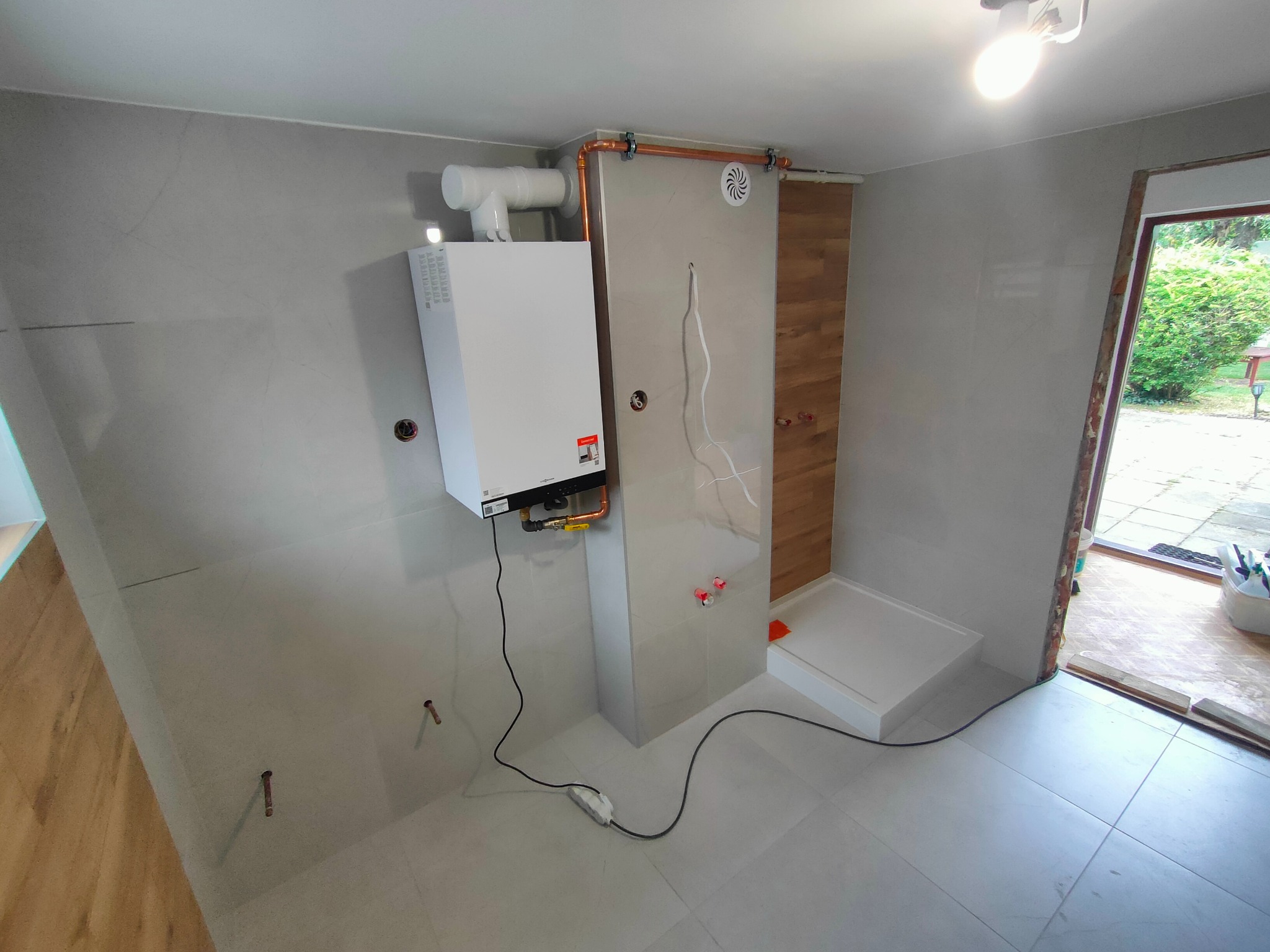 Nowoczesna łazienka w trakcie remontu: biały piec grzewczy na szarej, wykafelkowanej ścianie, obok kabina prysznicowa i okno z widokiem na zieleń.