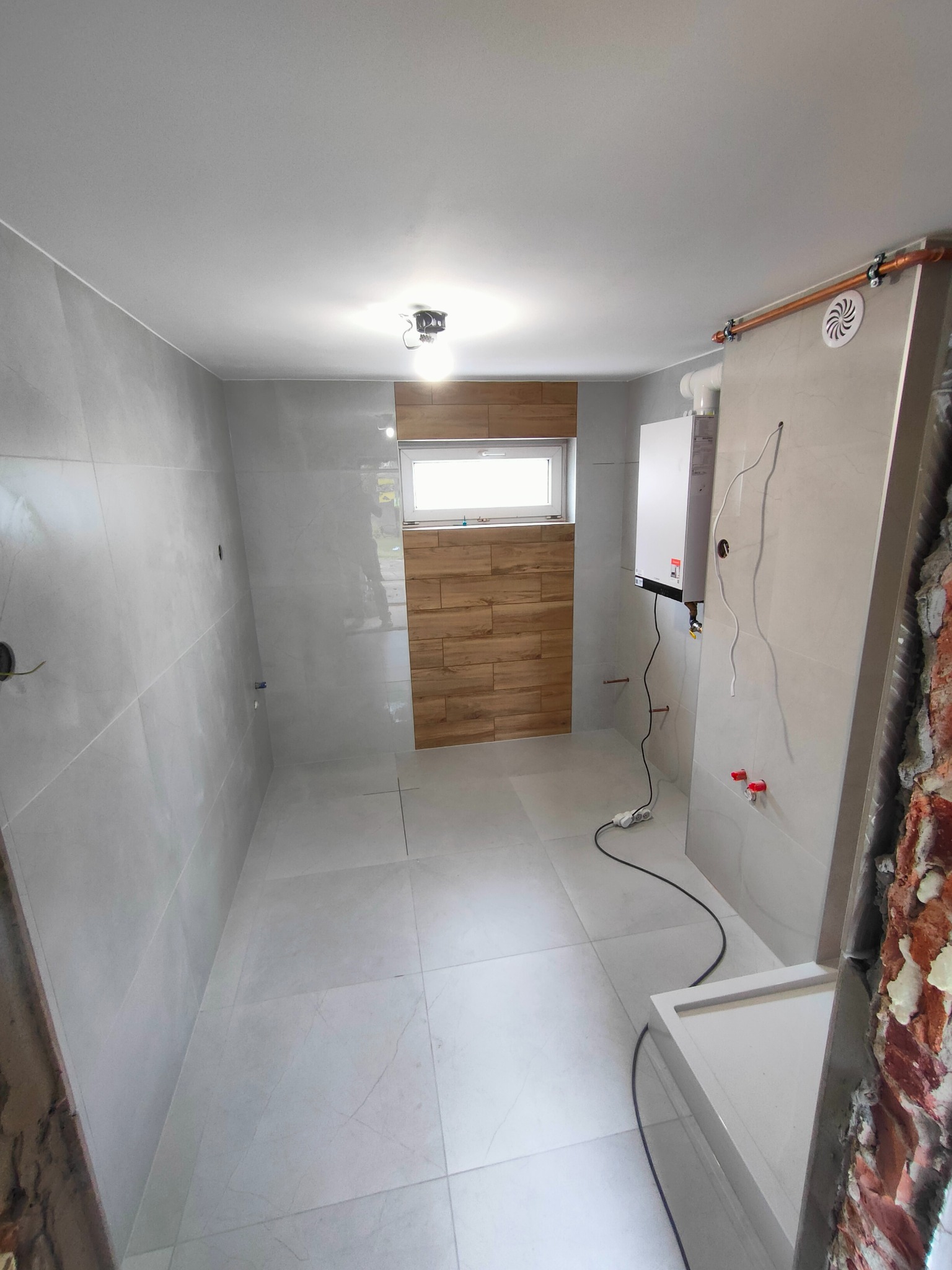 Wykończona łazienka z jasnymi płytkami, drewnopodobną ścianą i piecem grzewczym. Widoczne instalacje hydrauliczne i elektryczne. Częściowo wykończone ściany.