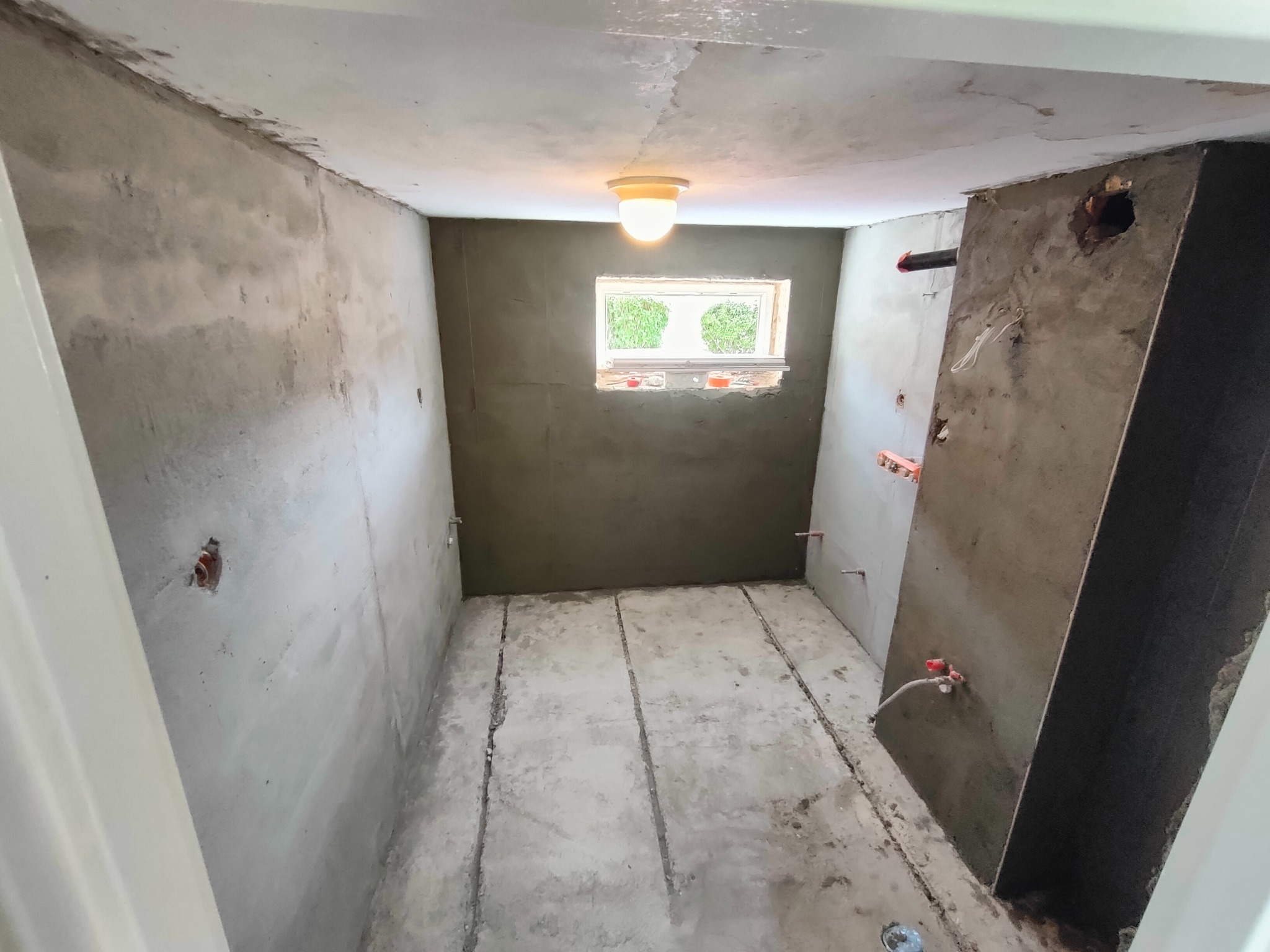 Surowy stan łazienki w trakcie remontu: ściany i podłoga w szarym tynku/betonie, widoczne instalacje, okno z zielenią na zewnątrz, sufit z lampą.