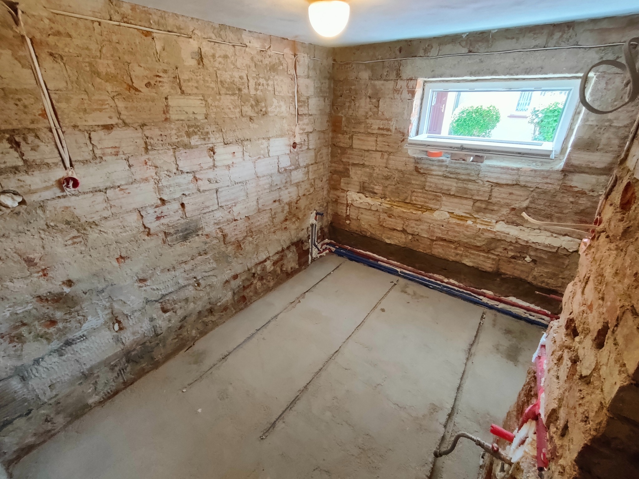 Surowa łazienka w trakcie remontu: odsłonięta cegła, instalacje podłogowe i okno z widokiem na zieleń. Etap prac przed wykończeniem, widoczne przewody i rury.