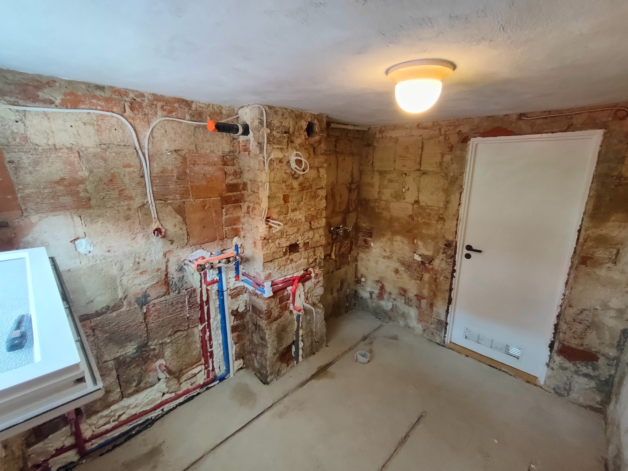 Surowa łazienka w trakcie remontu: odsłonięta cegła, nowe rury wodne (czerwone/niebieskie), białe kable i drzwi. Oświetlenie sufitowe. Widok na wnętrze przed wykończeniem.