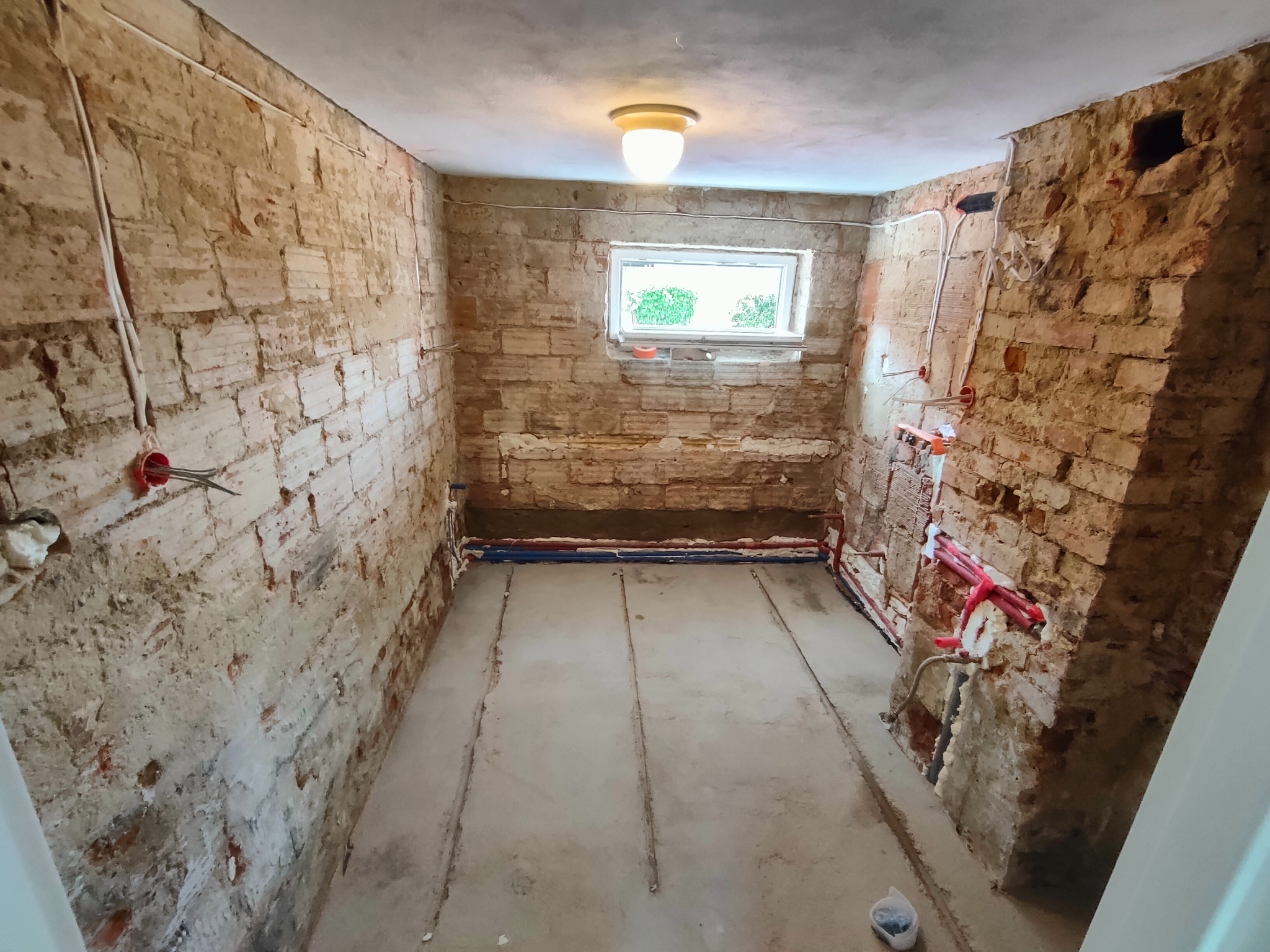 Łazienka w trakcie remontu: odkryte cegły, instalacje hydrauliczne w kolorze czerwonym i niebieskim, surowa podłoga, okno z widokiem na zieleń.