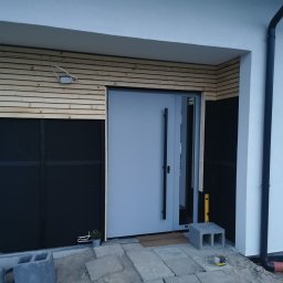 Rafał Pichnicki - Montaż drzwi wejściowych, widoczne drewniane panele elewacyjne w trakcie instalacji, czarna membrana, poziomica i betonowe bloczki na ziemi.