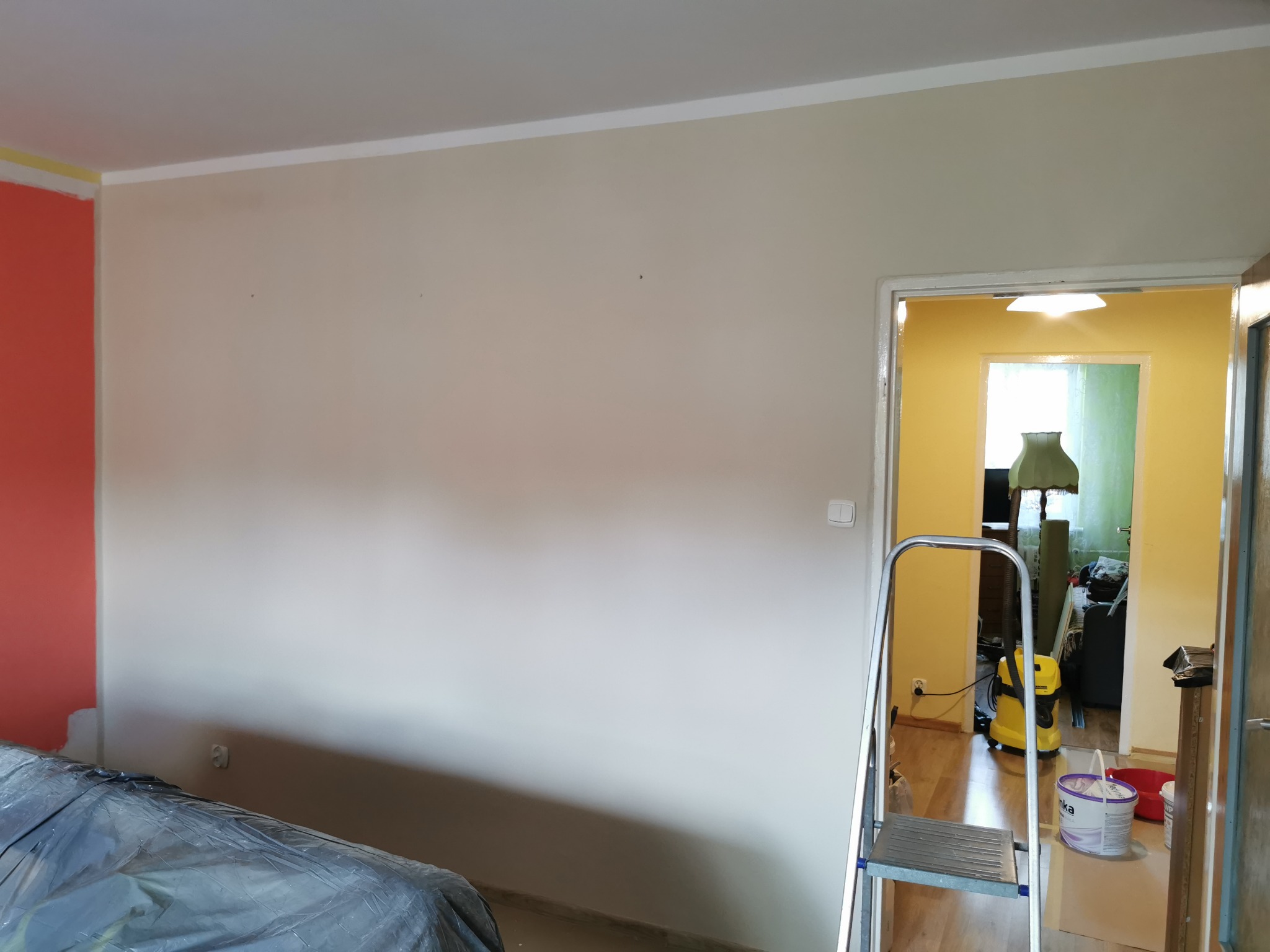 Pokój w trakcie malowania: ściana pomalowana częściowo na pomarańczowo, reszta na kremowo, zabezpieczone meble folią, drabina, wiaderko z farbą, odkurzacz w tle.