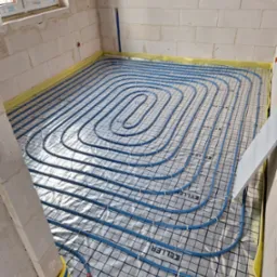 Niebieskie rury ogrzewania podłogowego ułożone na folii z nadrukiem 'KELLER' w surowym pomieszczeniu z białych bloczków, widoczne fragmenty czerwonych rur przy ścianie.