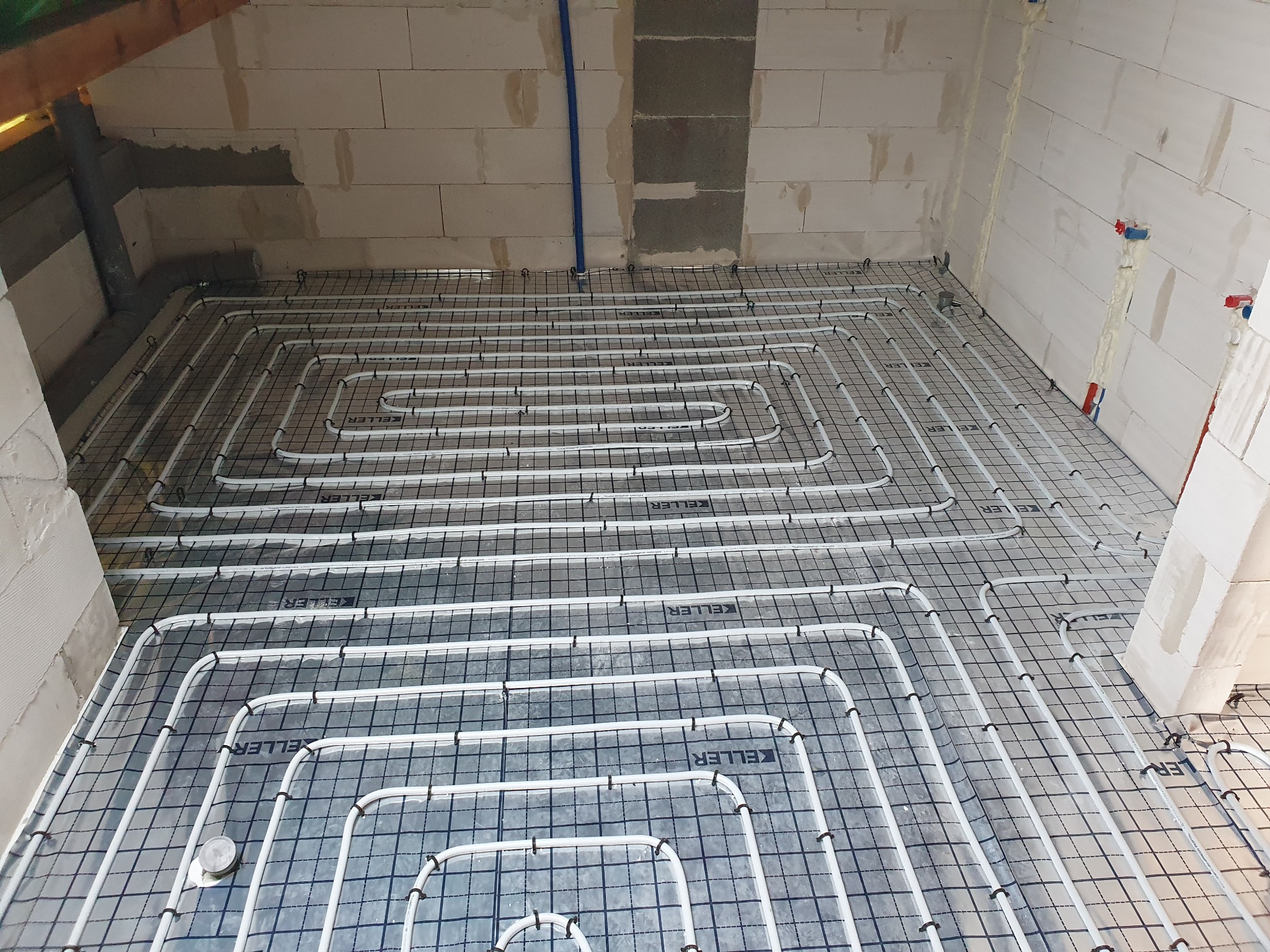 Białe rury ogrzewania podłogowego ułożone na czarnej siatce w surowym wnętrzu z białych bloczków.