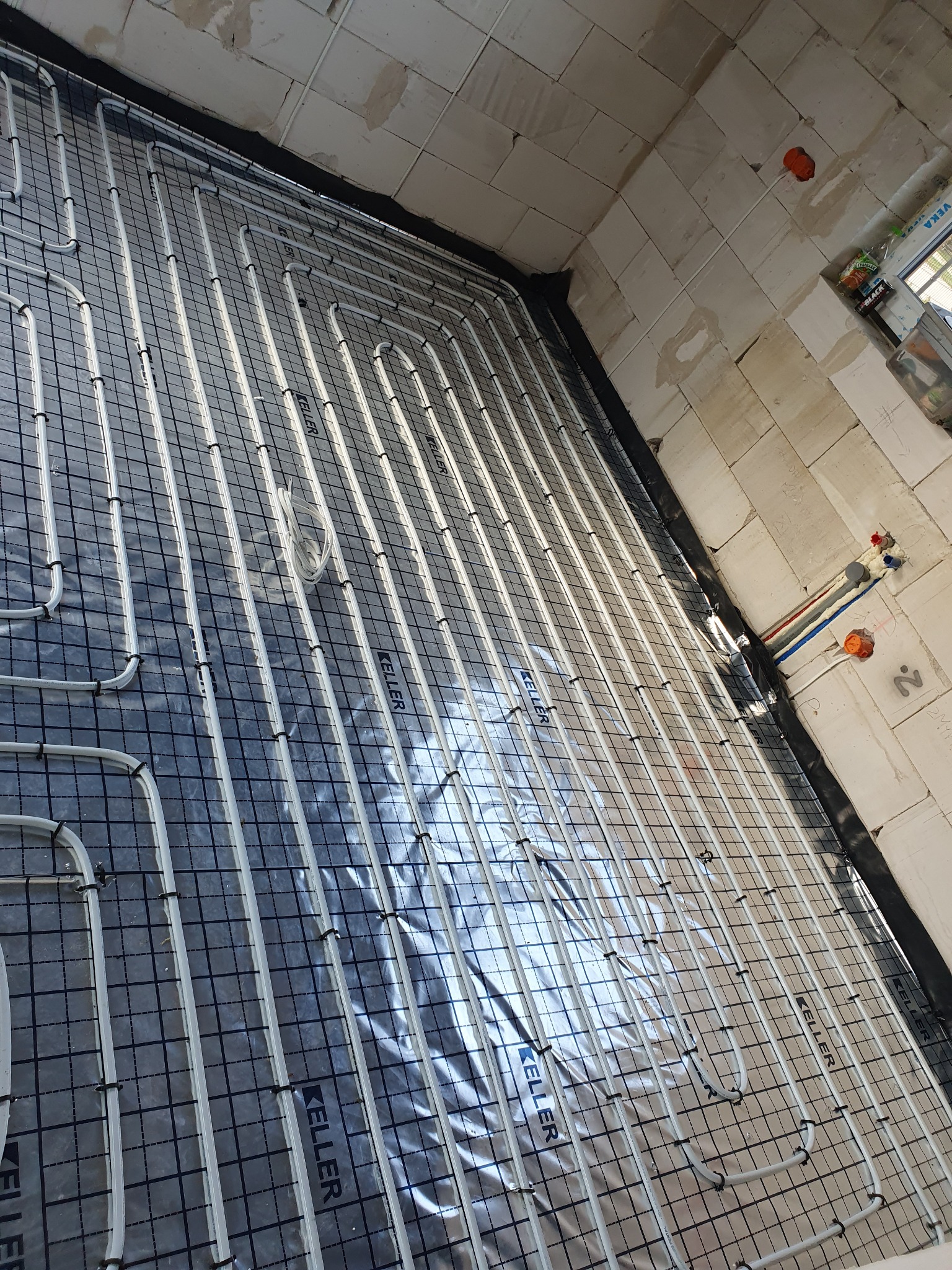 Białe rurki ogrzewania podłogowego ułożone na srebrnej folii z nadrukiem siatki i logo Keller, zamocowane czarnymi klipsami do siatki, widok z góry w surowym wnętrzu z bloczków.