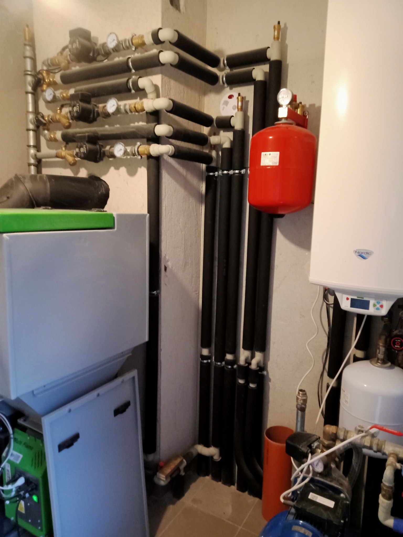 Kotłownia z piecem CO, instalacją grzewczą z rurami w izolacji, czerwonym zbiornikiem wyrównawczym, pompą obiegową i otwartą obudową kotła.