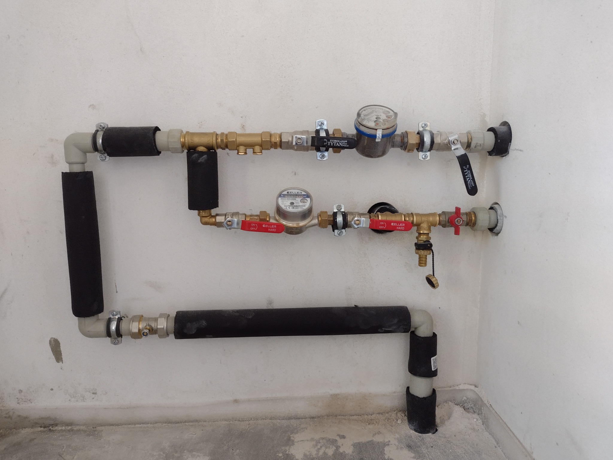 Dwa równoległe rzędy rur z licznikami wody, zaworami i izolacją termiczną, zamontowane przy białej ścianie w rogu pomieszczenia.