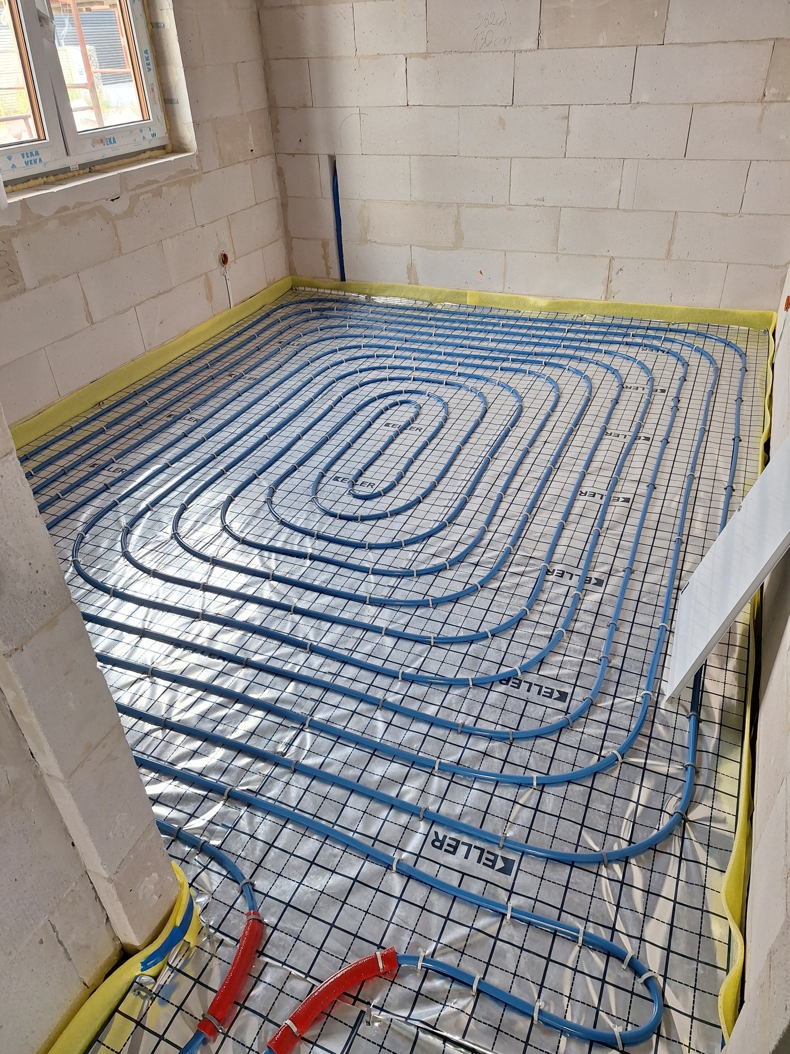 Niebieskie rury ogrzewania podłogowego ułożone na folii z nadrukiem 'KELLER' w surowym pomieszczeniu z białych bloczków, widoczne fragmenty czerwonych rur przy ścianie.