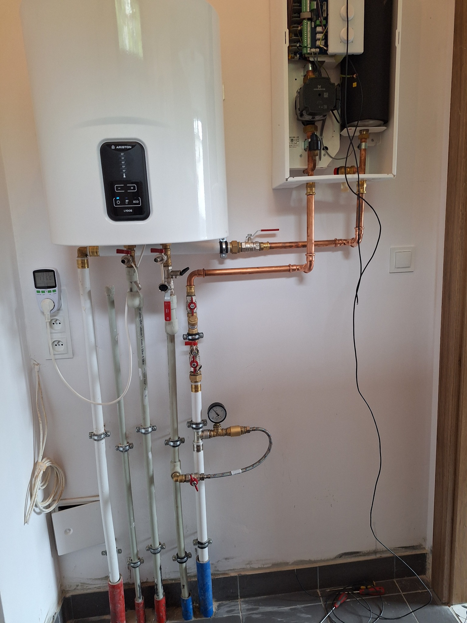 Biały bojler Ariston z elektronicznym panelem sterowania, podłączony miedzianymi rurami do instalacji grzewczej z zaworami, manometrem i pompką obiegową w białej obudowie, zamontowany na ścianie...