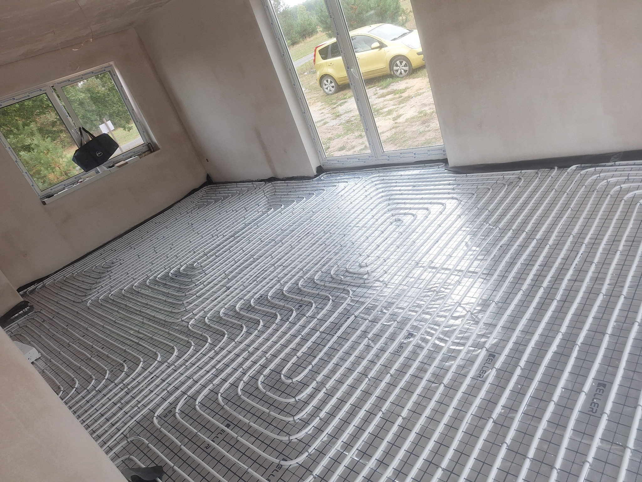 Białe rury ogrzewania podłogowego ułożone na czarnej macie w surowym pomieszczeniu. Przez okno widoczny żółty samochód. Torba leży na parapecie.