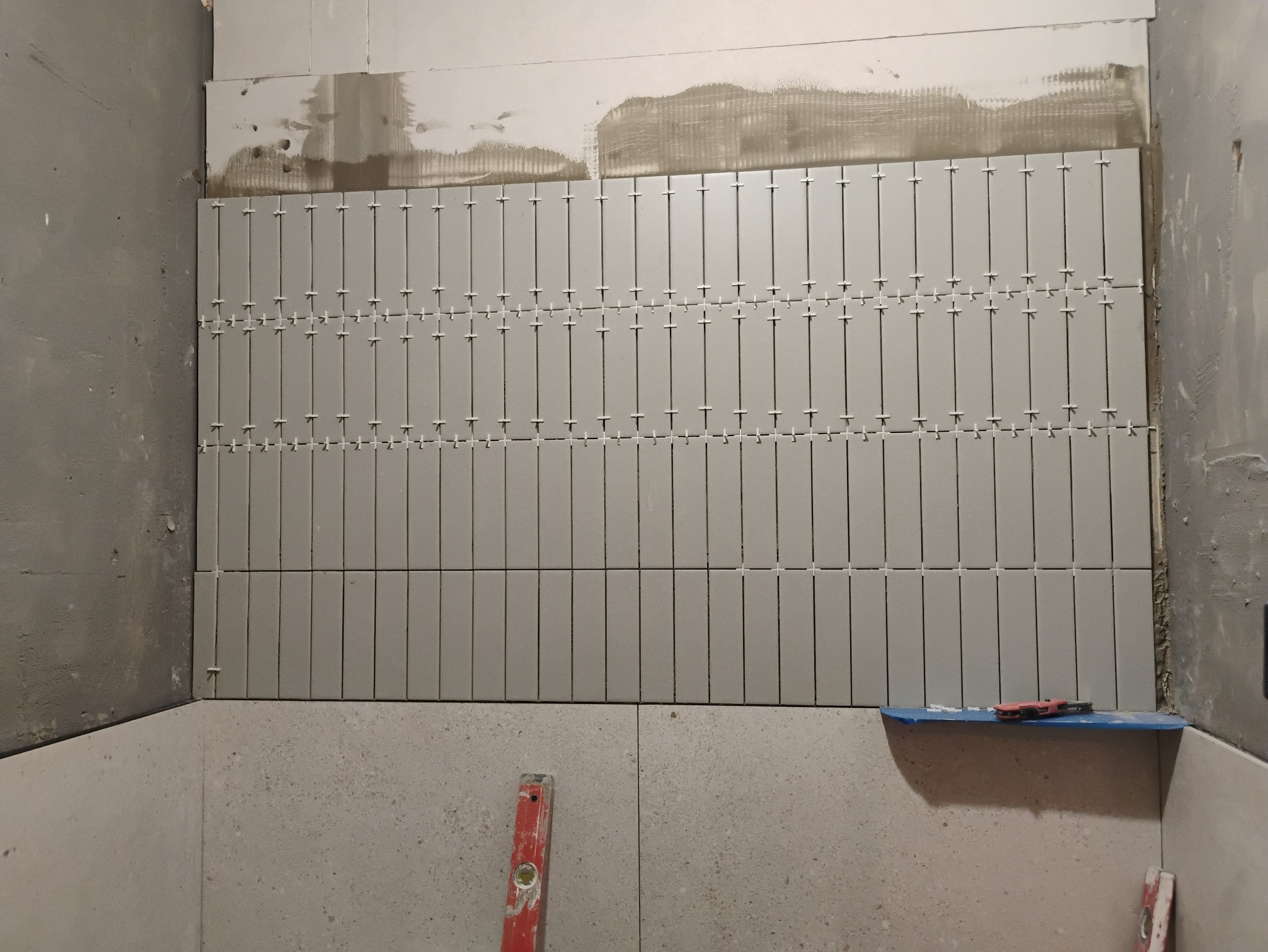 Świeżo ułożone, pionowe, szare płytki na ścianie z białymi krzyżykami dystansowymi, widoczny poziomica i nieukończona ściana obok.