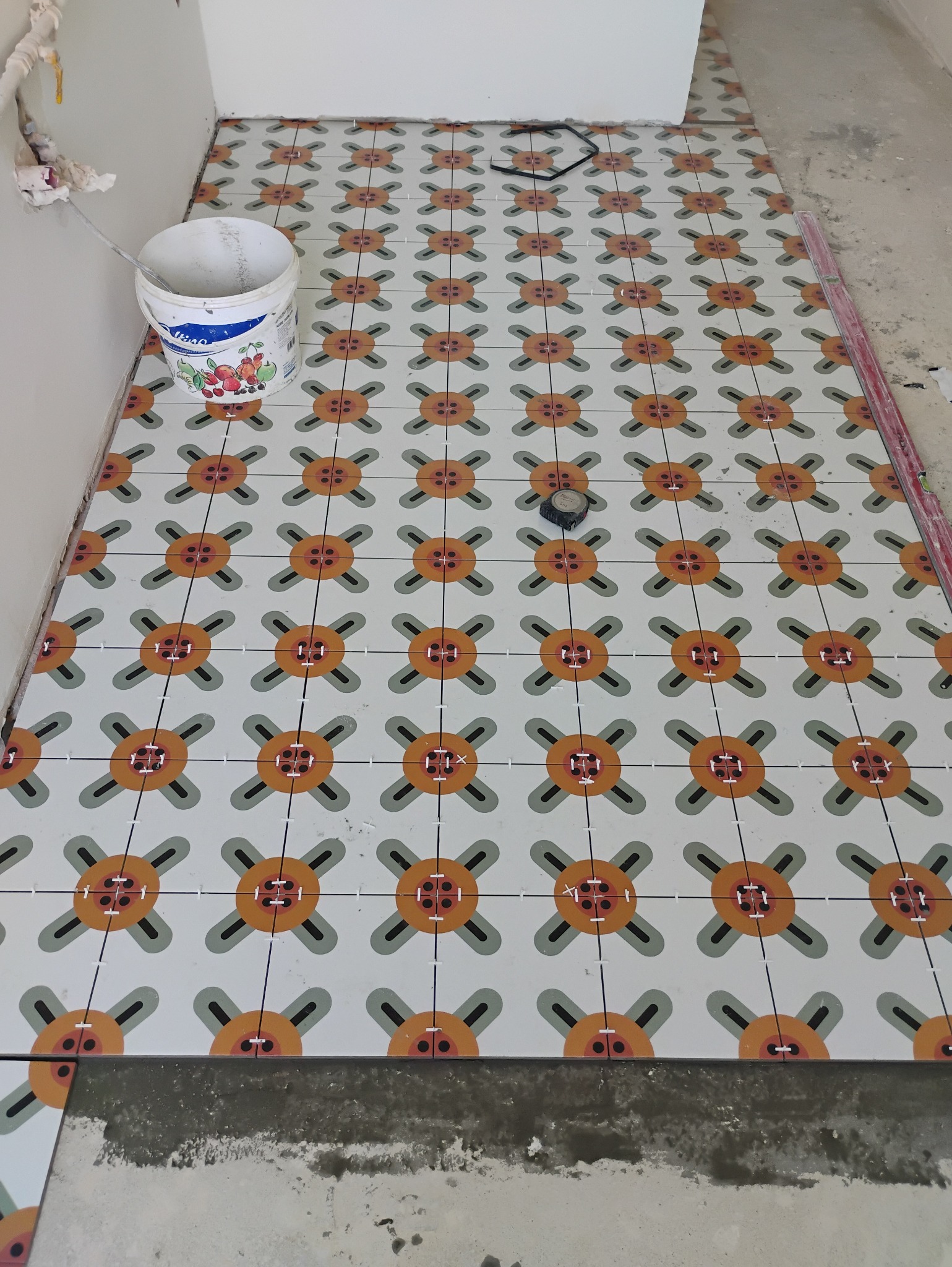 Układanie wzorzystych płytek podłogowych w trakcie prac wykończeniowych, widoczny pojemnik z farbą i krzyżyki dystansowe.