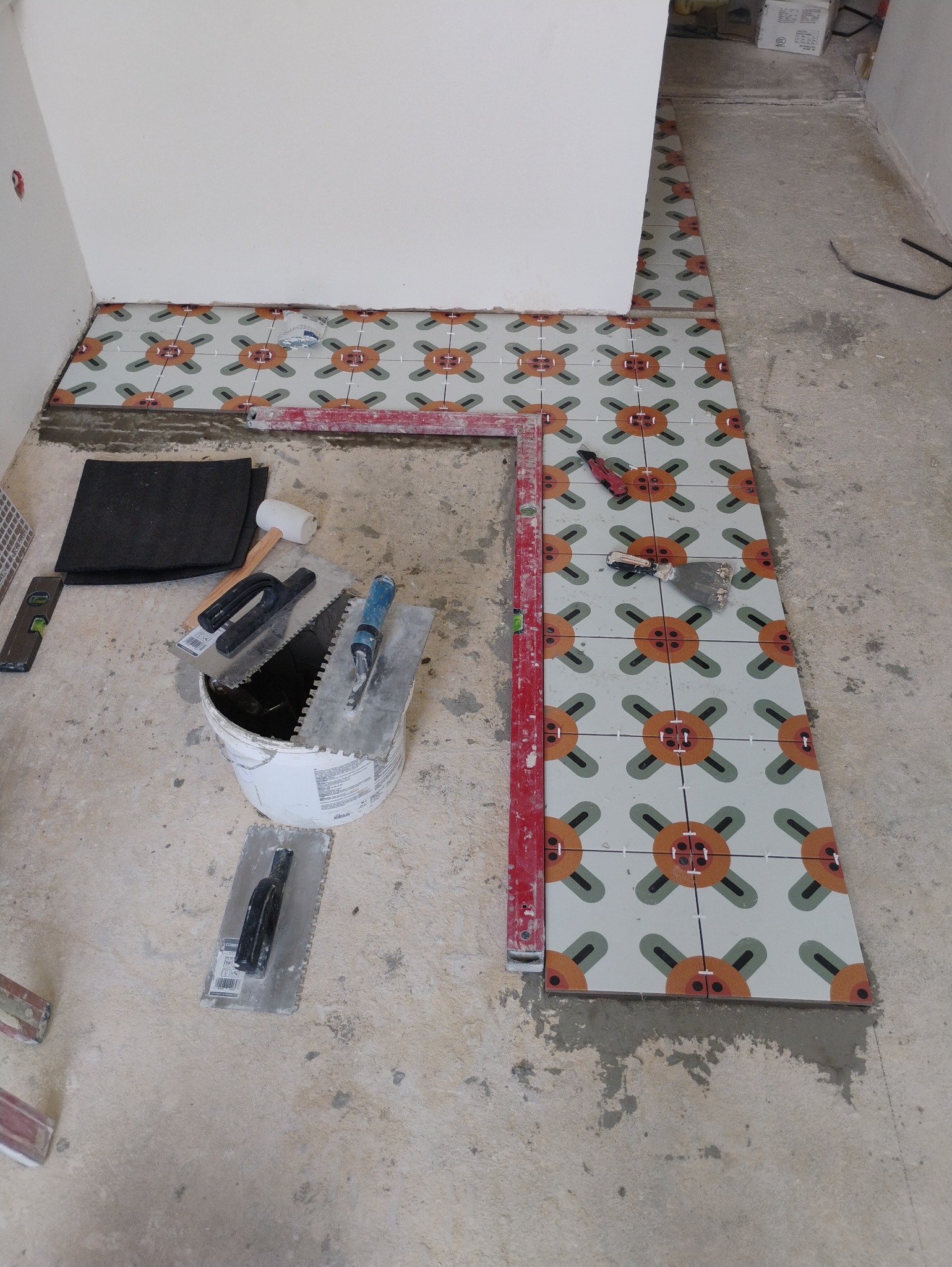 Układanie wzorzystych płytek podłogowych w pomieszczeniu, widoczne narzędzia glazurnicze, poziomica i wiadro z klejem.