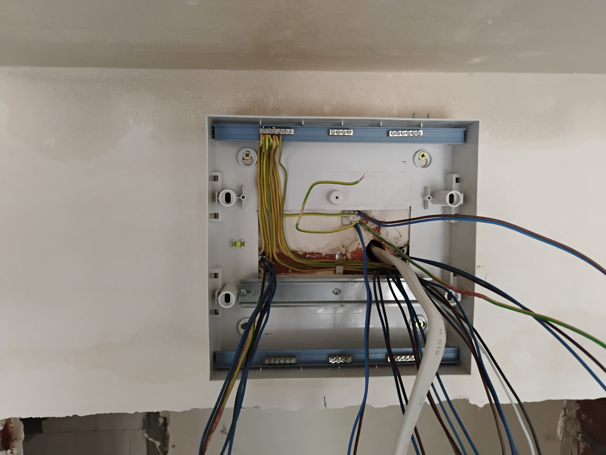 Otwarta skrzynka elektryczna w ścianie z widocznymi przewodami różnokolorowymi i listwami zaciskowymi.