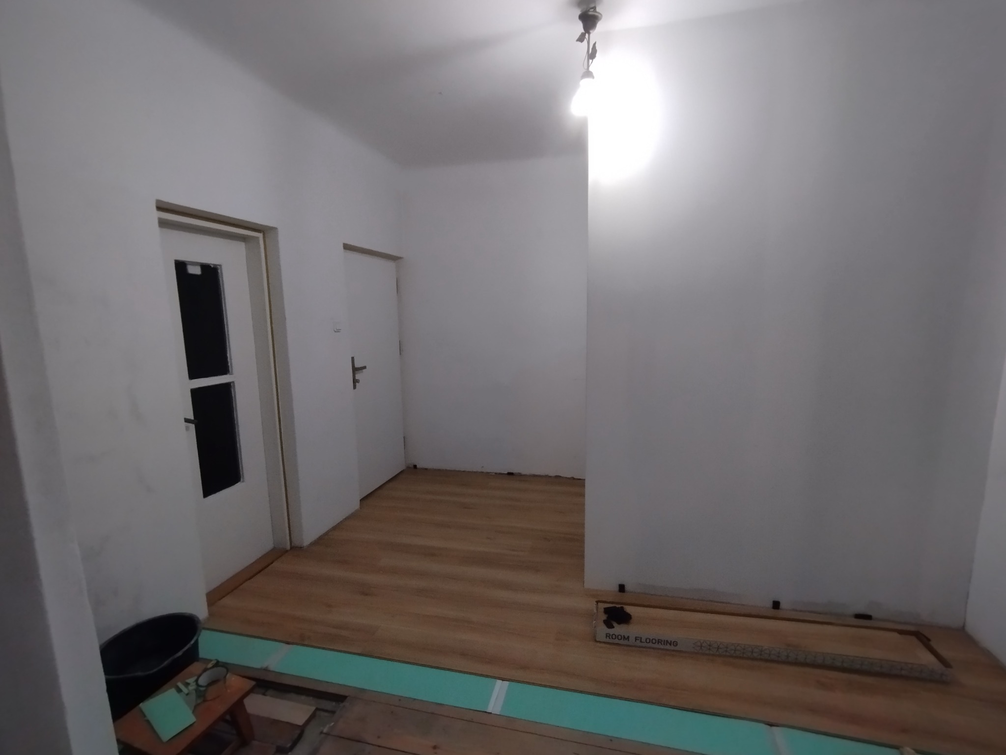 Pokój w trakcie układania paneli podłogowych, widoczne płyty izolacyjne, częściowo ułożone panele, opakowanie paneli i narzędzia.