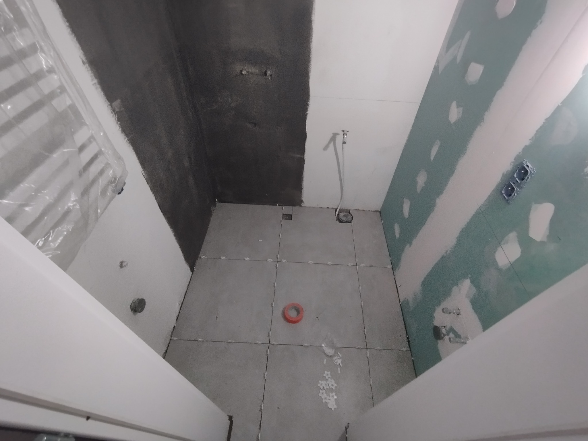 Wnętrze łazienki w trakcie prac wykończeniowych: szpachlowane ściany, częściowo położone szare płytki podłogowe z krzyżykami dystansowymi, widoczne instalacje hydrauliczne i elektryczne.