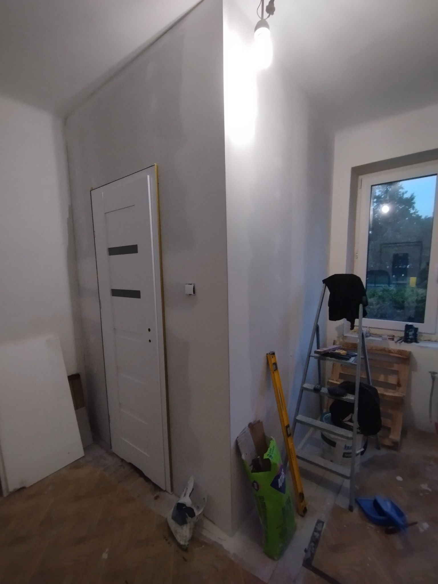 Wnętrze w trakcie remontu: białe ściany po szpachlowaniu, białe drzwi z poziomymi przeszkleniami, drabina aluminiowa, poziomica i worek zaprawy Atlas.