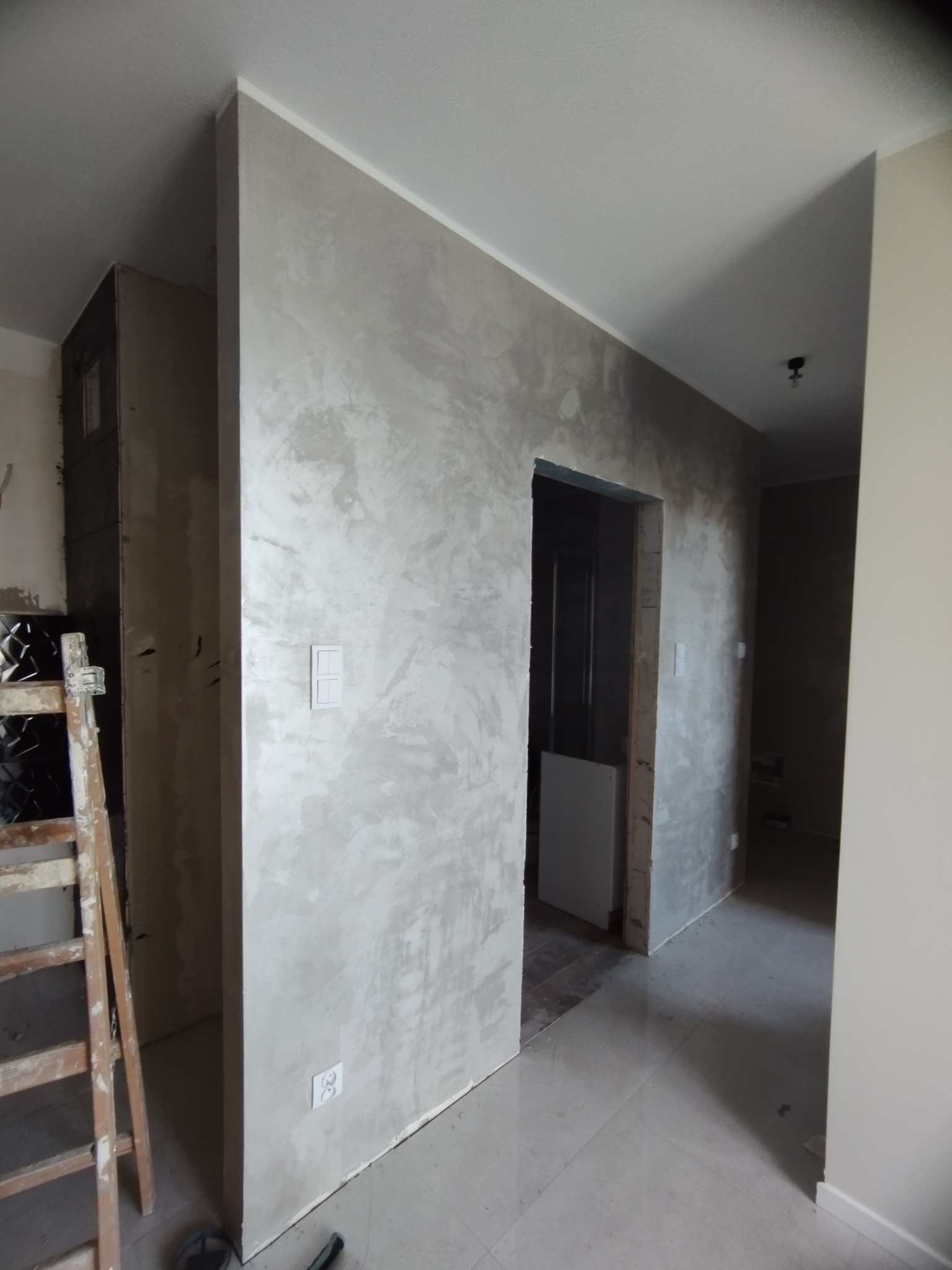 Ściana w trakcie szpachlowania i malowania na szaro z widoczną fakturą, fragment drabiny malarskiej i gniazdka elektryczne, widok na sąsiednie pomieszczenie.