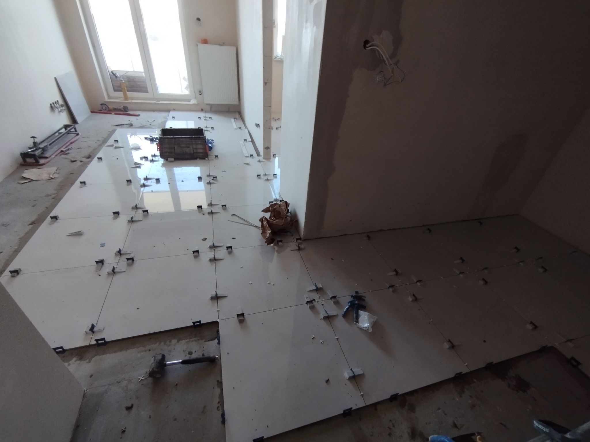 Wnętrze w trakcie układania płytek podłogowych, widoczne narzędzia glazurnicze, krzyżyki dystansowe, poziomica i fragmenty niezabezpieczonych przewodów elektrycznych.