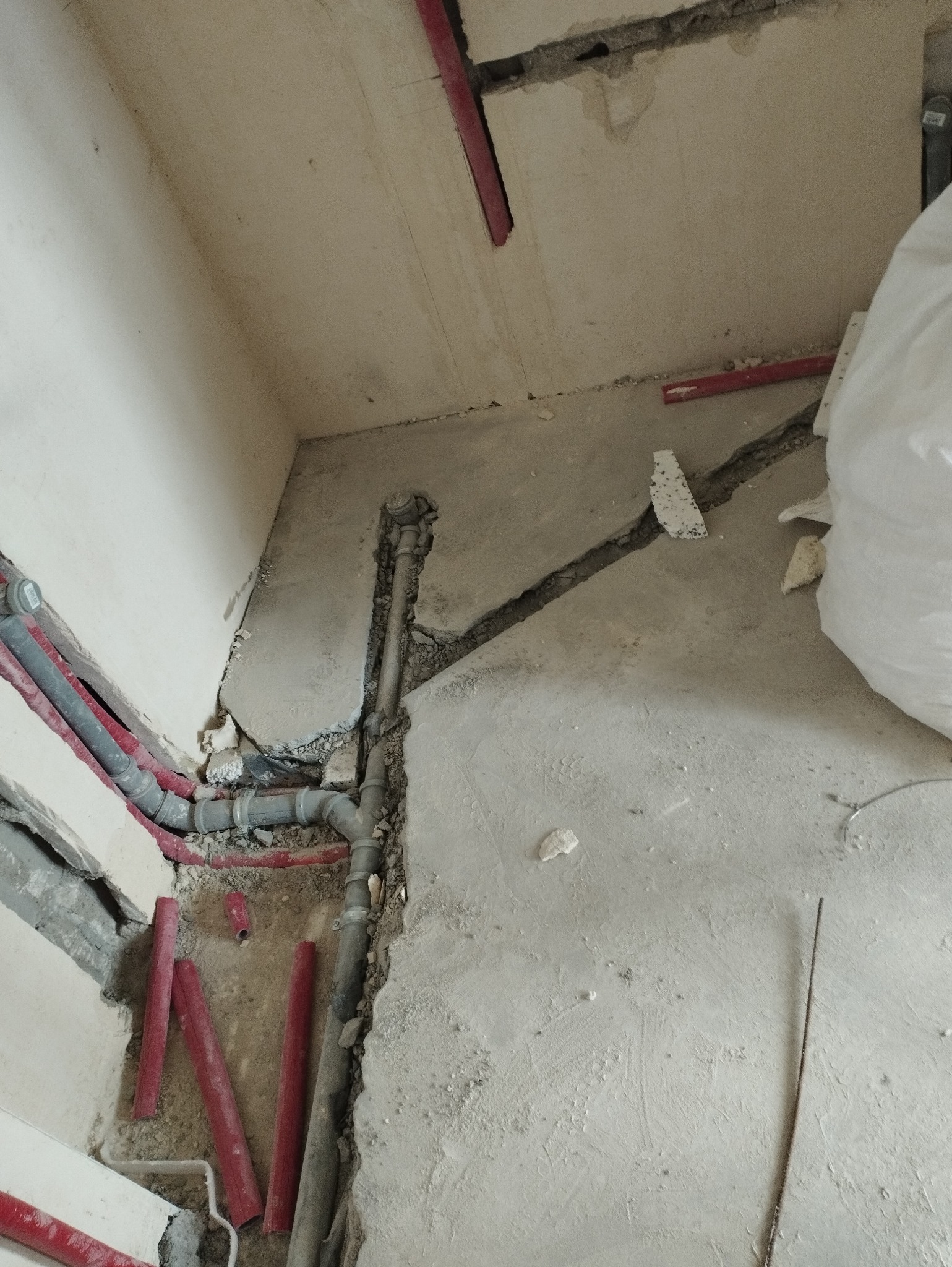 Fragment wnętrza w trakcie prac instalacyjnych z widocznymi rurami grzewczymi, kanalizacyjnymi i fragmentem ściany z wykuciami, surowy stan wykończenia.