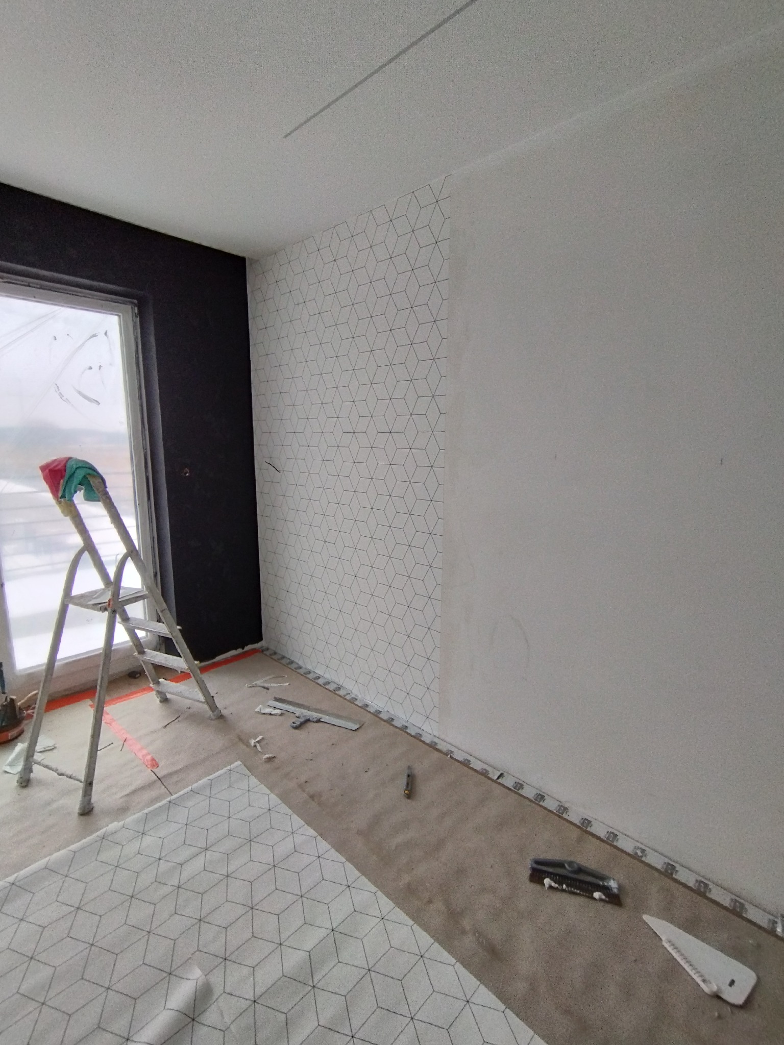 Pokój w trakcie tapetowania: fragment ściany z geometrycznym wzorem, drabina z zieloną szmatką, narzędzia do tapetowania na podłodze zabezpieczonej folią.