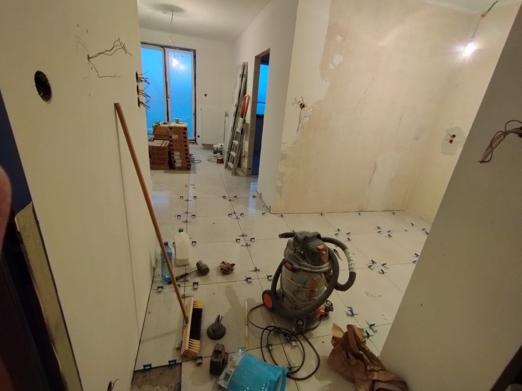 Wnętrze w trakcie układania płytek podłogowych, widoczne krzyżyki dystansowe, odkurzacz przemysłowy, szpachlowana ściana z wystającymi przewodami elektrycznymi i przygotowane do montażu drzwi...