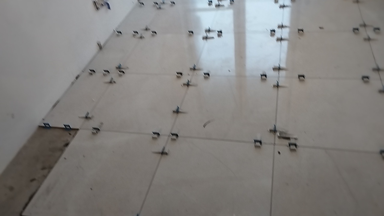 Ułożone, jasne płytki podłogowe z niebieskimi krzyżykami dystansowymi, widoczne niedociągnięcia przy ścianie.