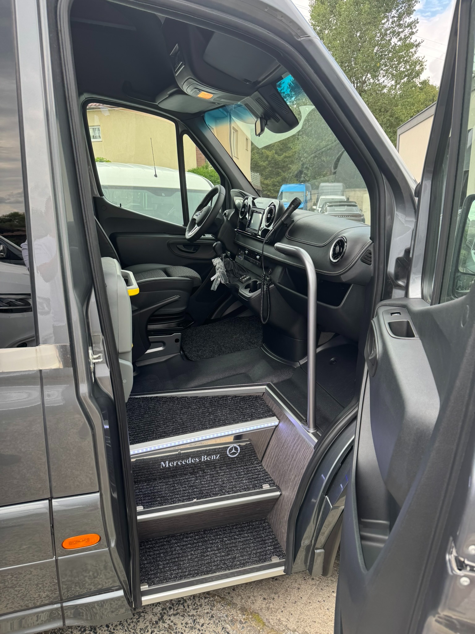 Wnętrze szarego busa Mercedes-Benz z otwartymi drzwiami, widoczne podświetlane stopnie z logo marki i poręczą.