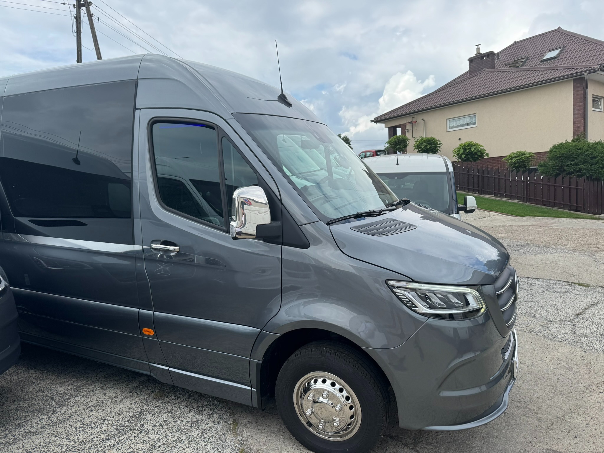 Szary bus pasażerski Mercedes-Benz Sprinter z chromowanymi lusterkami i kołpakami, stojący na parkingu przed beżowym domem z brązowym dachem.