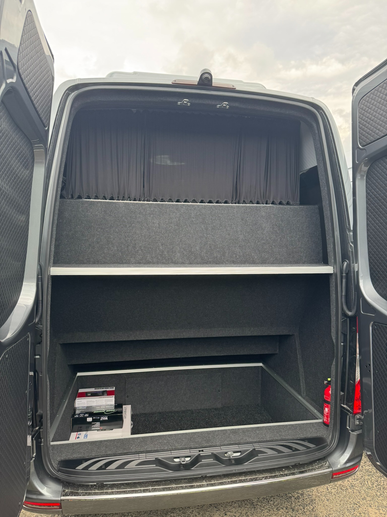 Wnętrze bagażnika dostawczego busa z półkami i zasłonami, widok z tyłu, puste półki z szarą wykładziną, na dolnej półce leżą pudełka z elektroniką.