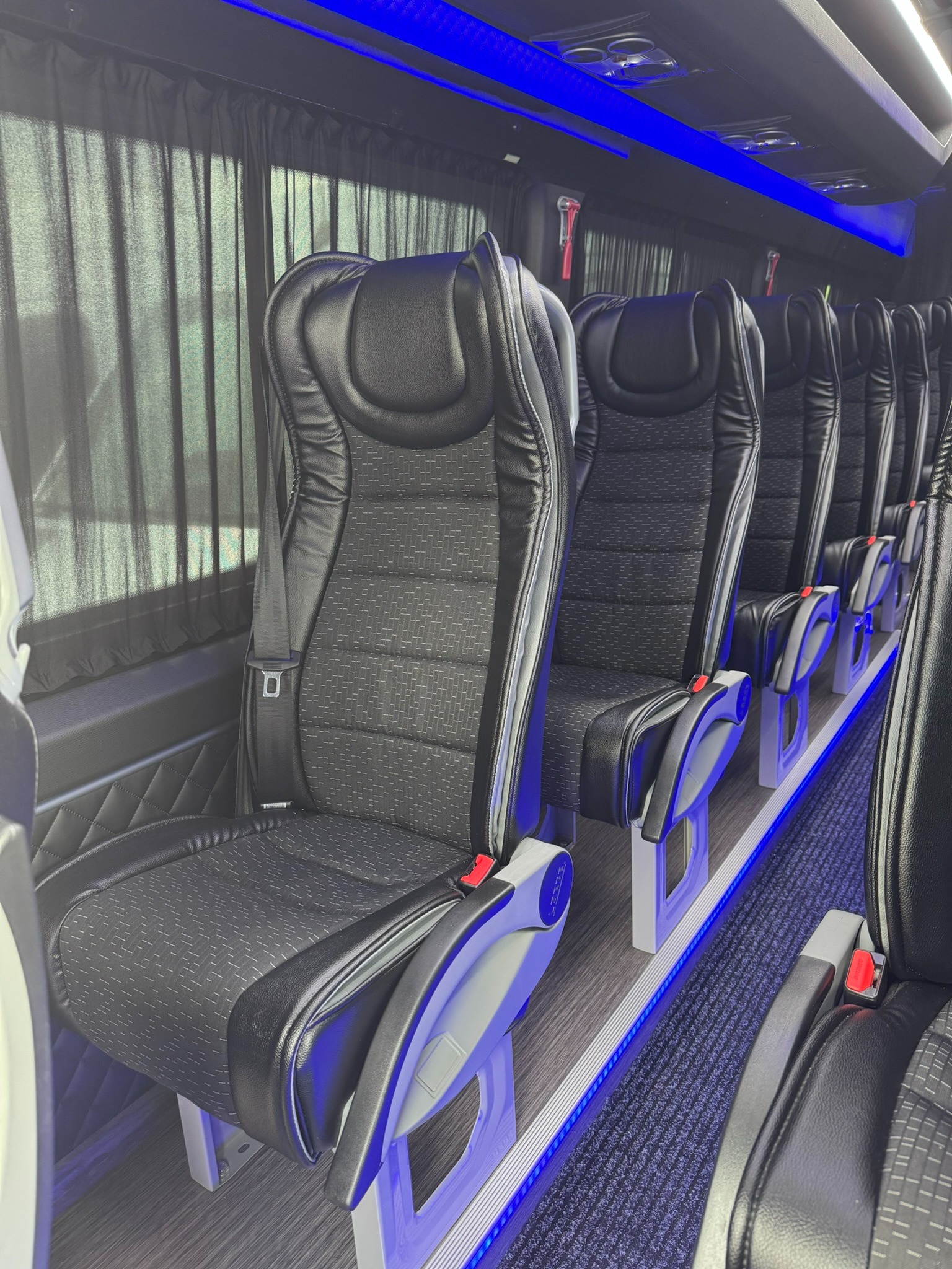 Wnętrze autobusu z rzędem czarnych, skórzanych foteli z pasami bezpieczeństwa, oświetlonych niebieskim światłem LED.