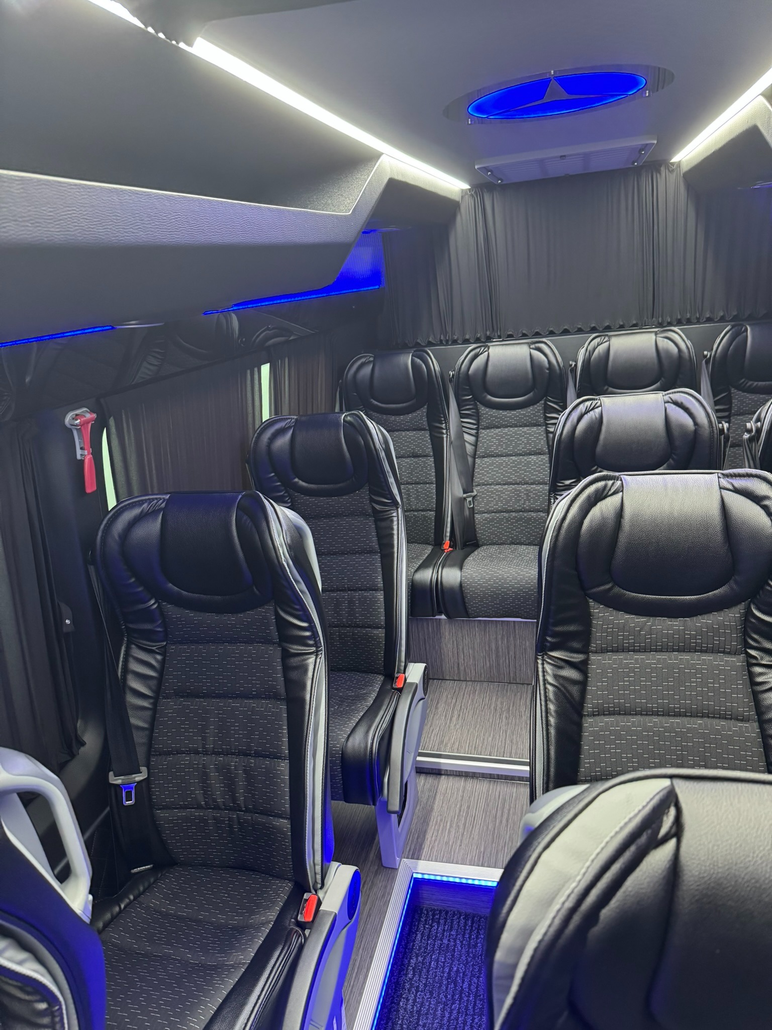 Wnętrze luksusowego busa z czarnymi skórzanymi fotelami, oświetleniem LED i zasłonami w oknach.
