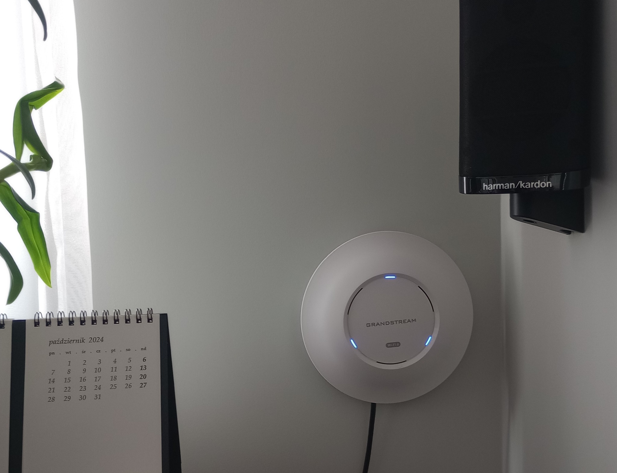 Biały punkt dostępowy Grandstream Wi-Fi 6 zamontowany na ścianie, obok głośnika Harman Kardon i kalendarza na październik 2024, z widoczną rośliną w tle.