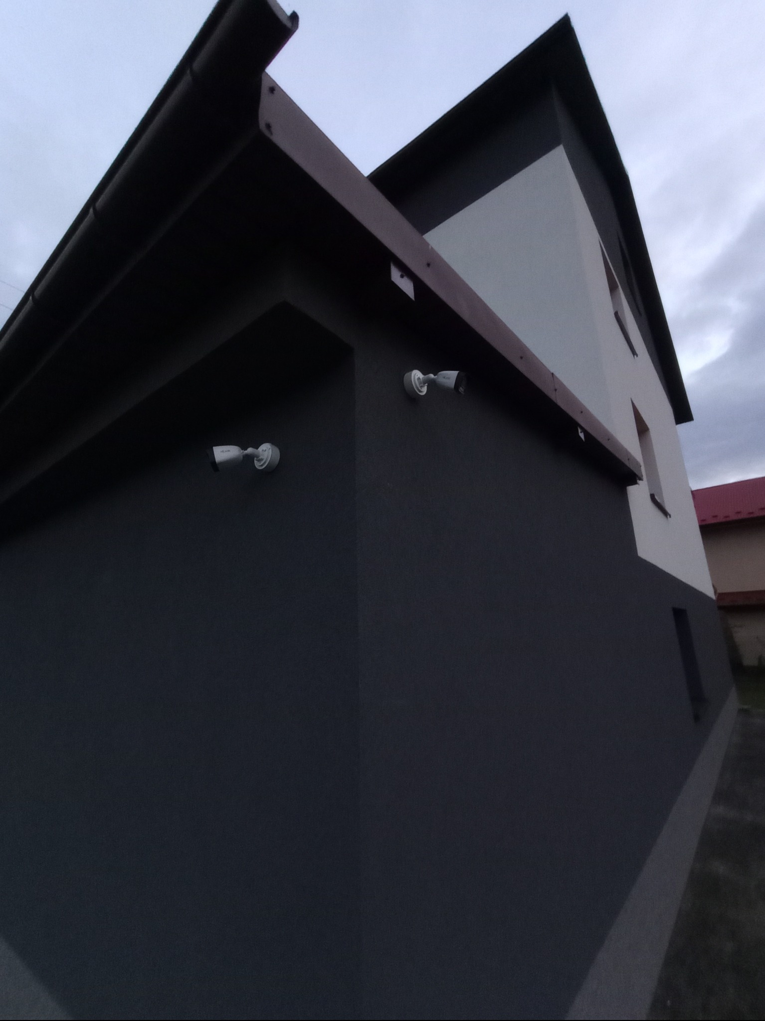 Zewnętrzny widok narożnika budynku z zainstalowanymi dwiema kamerami monitoringu na ciemnoszarej ścianie, widoczne elementy dachu i okna.