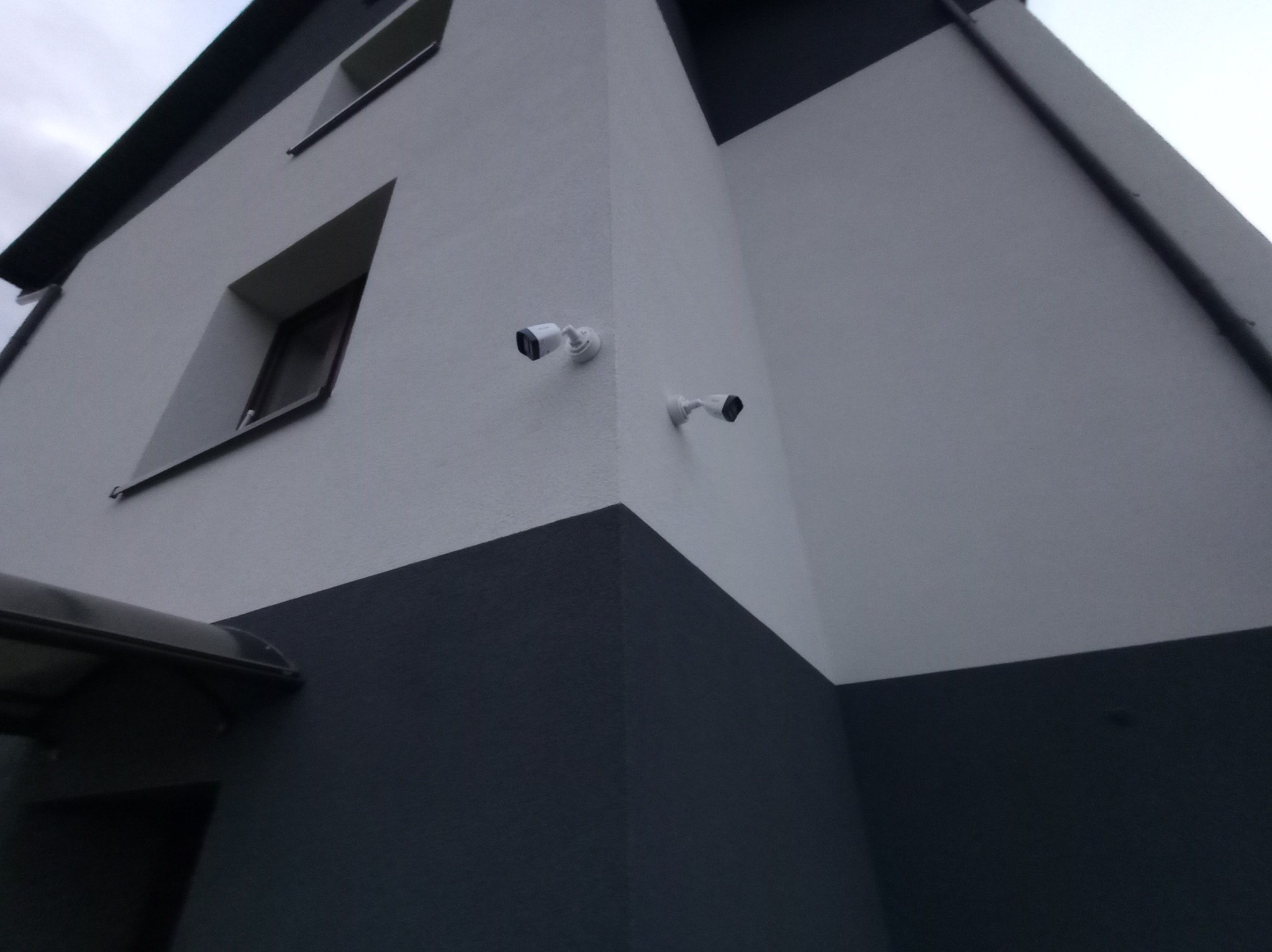 Dwie białe kamery monitoringu zamontowane na elewacji dwukondygnacyjnego budynku pomalowanego w odcieniach szarości, widoczne z dołu pod kątem.