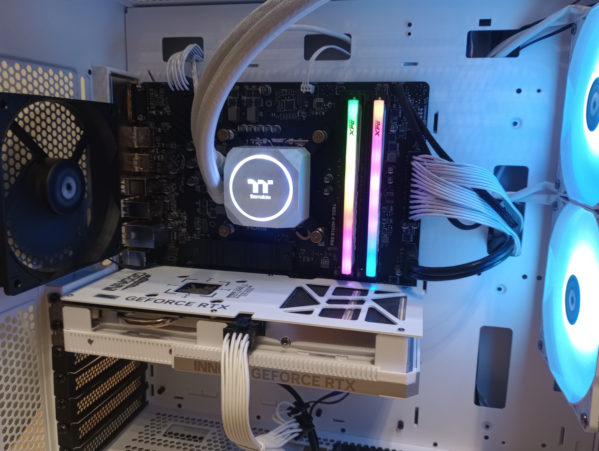 Wnętrze białej obudowy komputera stacjonarnego z podświetlanymi elementami: chłodzeniem wodnym Thermaltake, kartą graficzną GeForce RTX Inno3D i pamięcią RAM XPG z tęczowym podświetleniem.