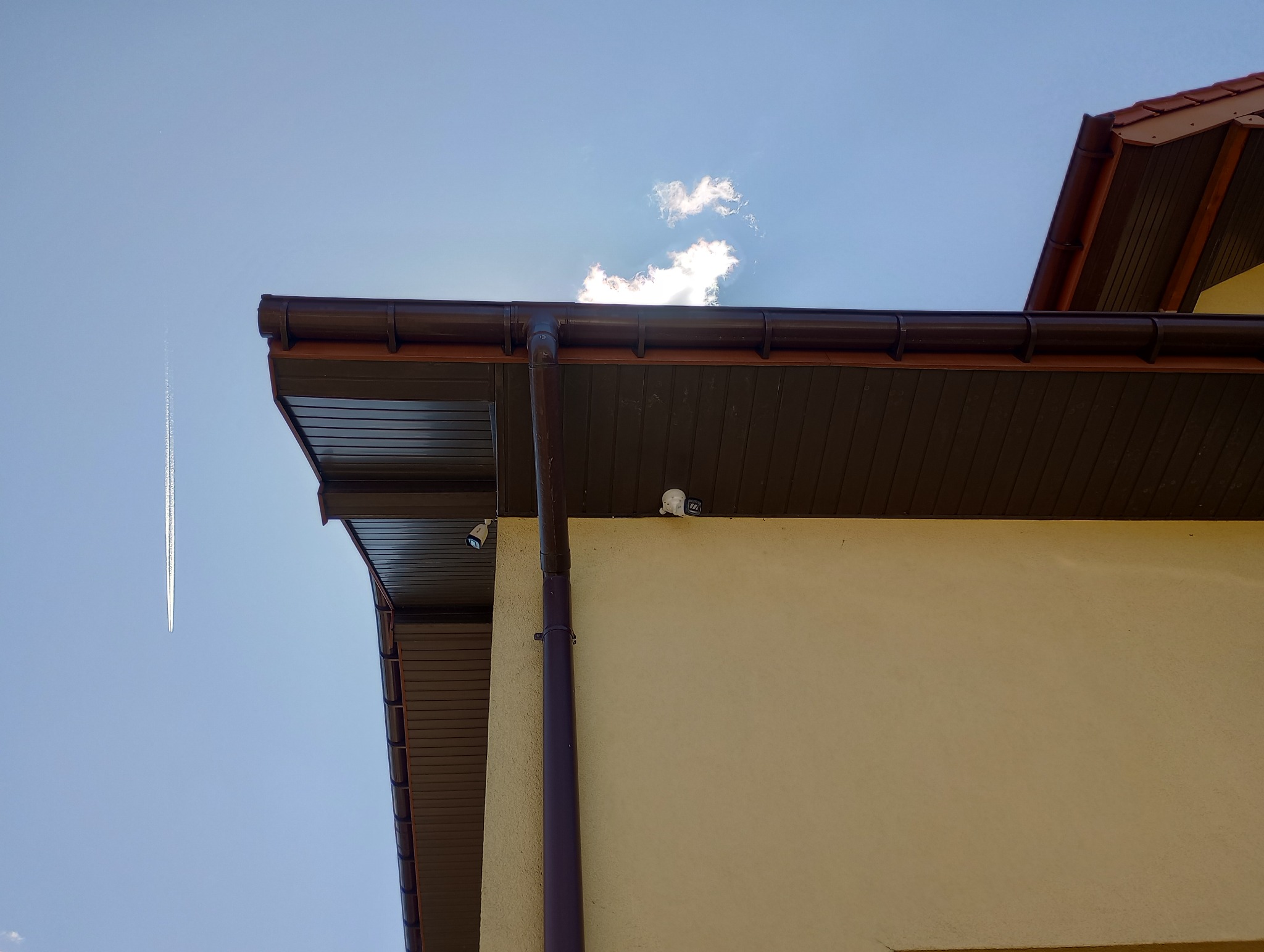 Kamera monitoringu zamontowana pod brązowym okapem budynku z beżową elewacją, na tle błękitnego nieba z smugą kondensacyjną samolotu.