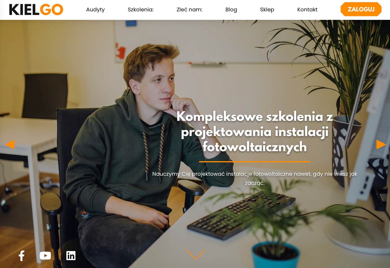 Młody mężczyzna w bluzie z kapturem siedzi przy biurku z klawiaturą i rośliną w doniczce, przeglądając stronę internetową kielgo.pl dotyczącą szkoleń z projektowania instalacji fotowoltaicznych...